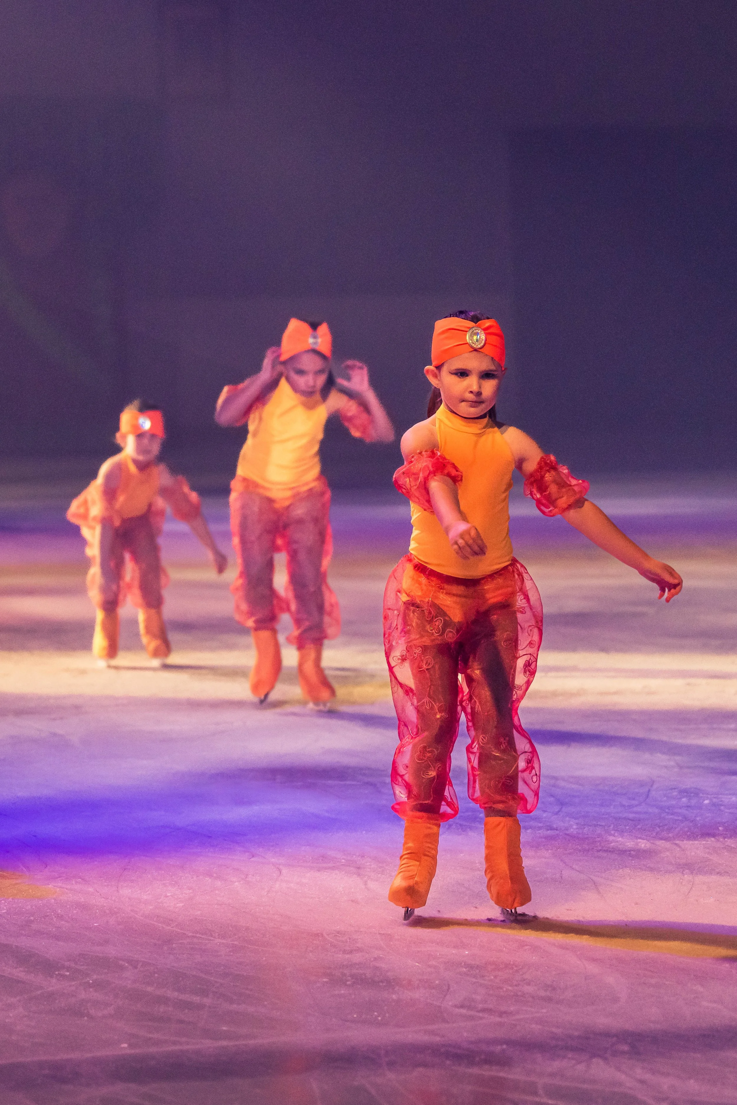 Four children practicing ice skating on a rink while wearing bright orange costumes with red and yellow accents, and orange headbands. The background is dark and the ice surface reflects purple and pink lighting.
