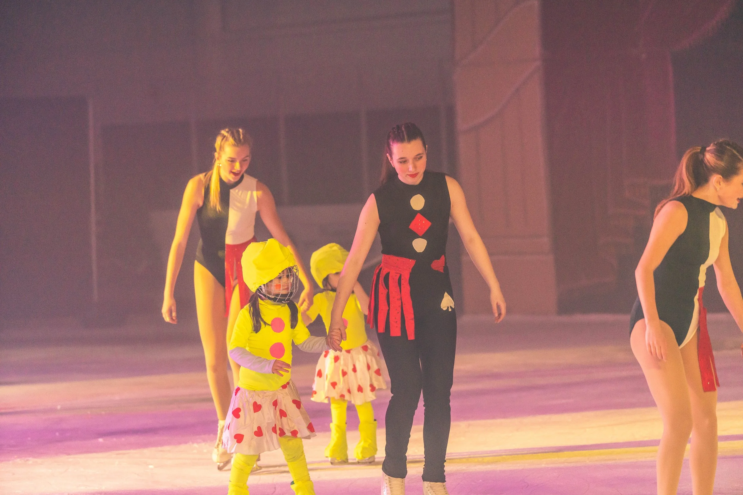 A group of young women and children in colorful costumes, holding hands, preparing for a performance on an ice rink.
