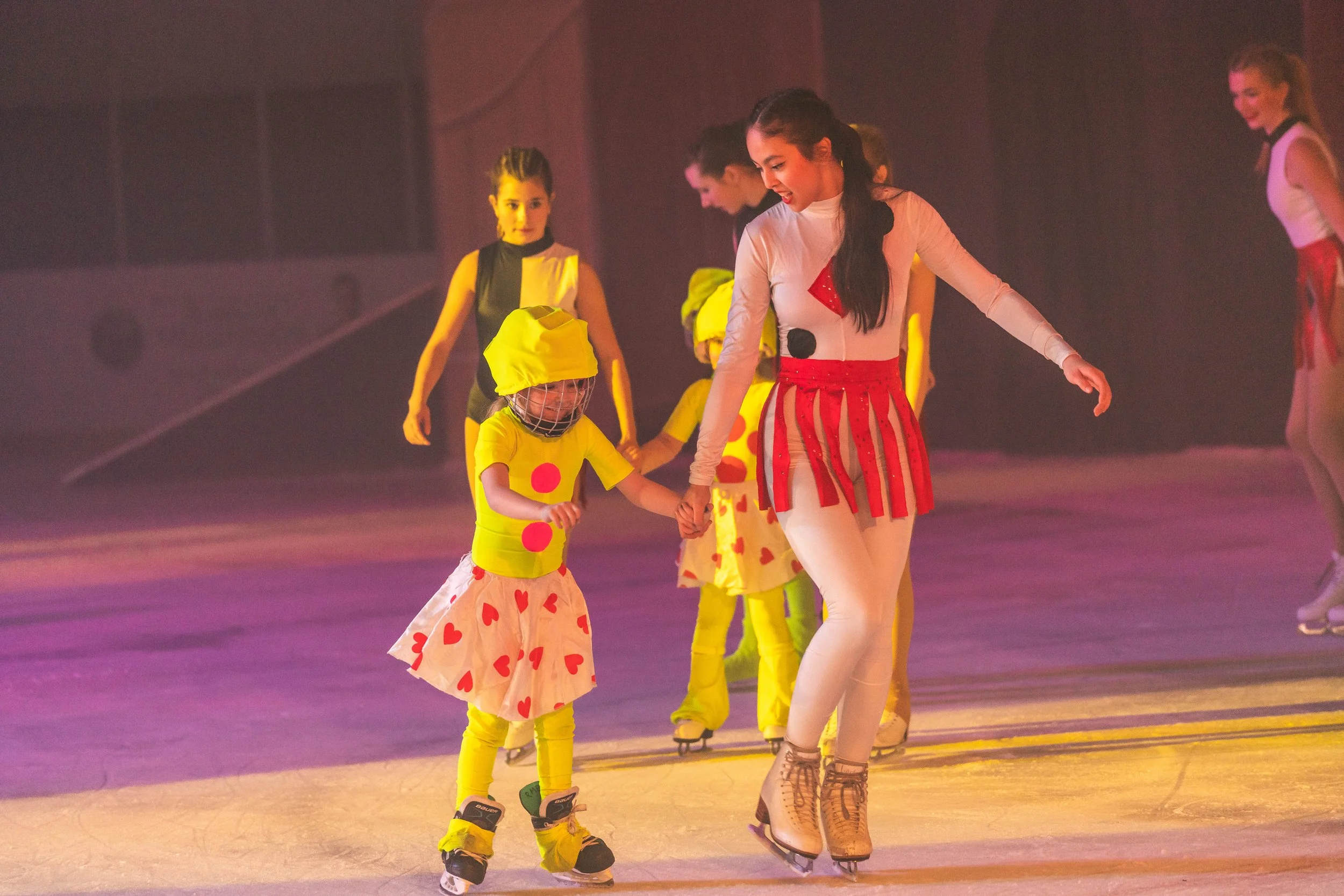 Children figure skating in colorful costumes on an ice rink, with a smiling young woman in a striped dress helping a girl dressed as a clown.