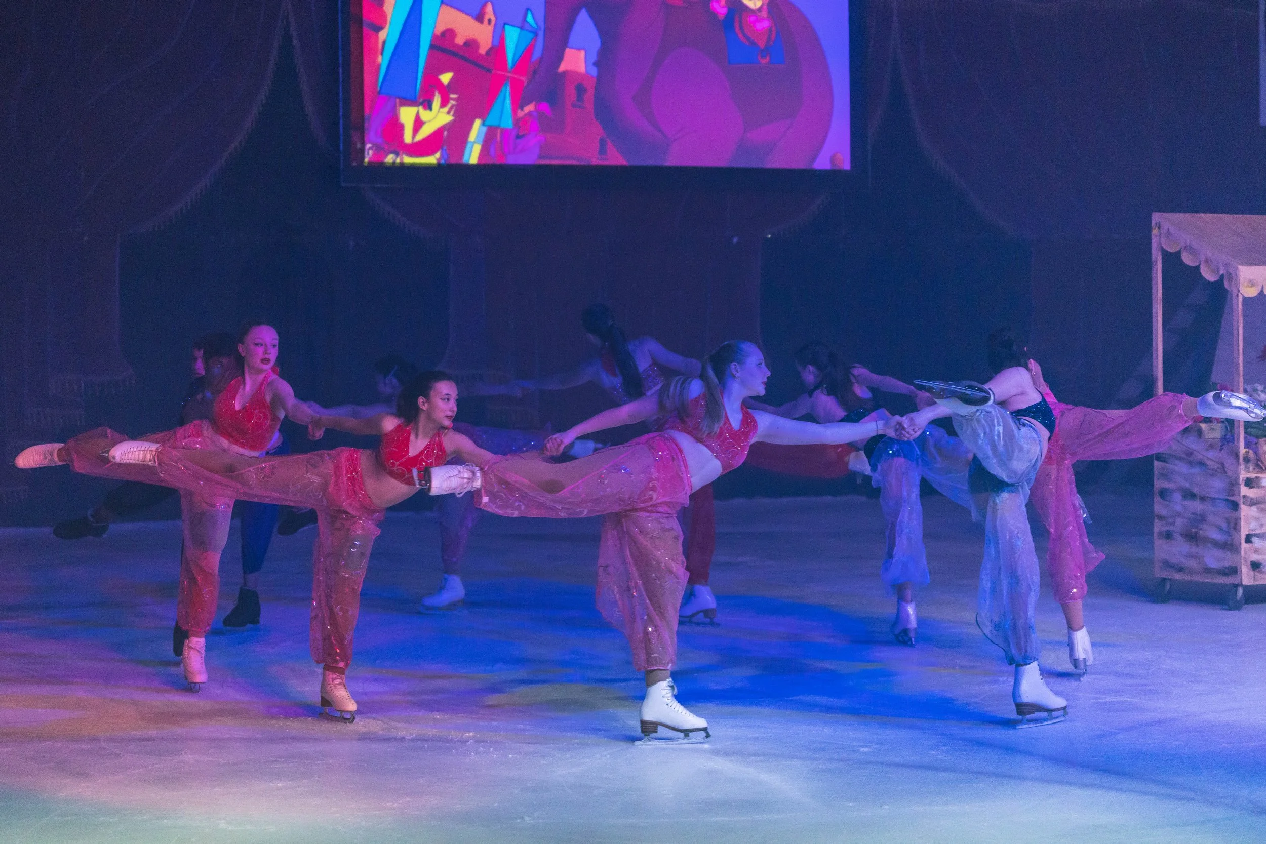 A group of female figure skaters performing a synchronized routine on ice, wearing colorful costumes, under dim stage lighting with a large animated screen in the background.