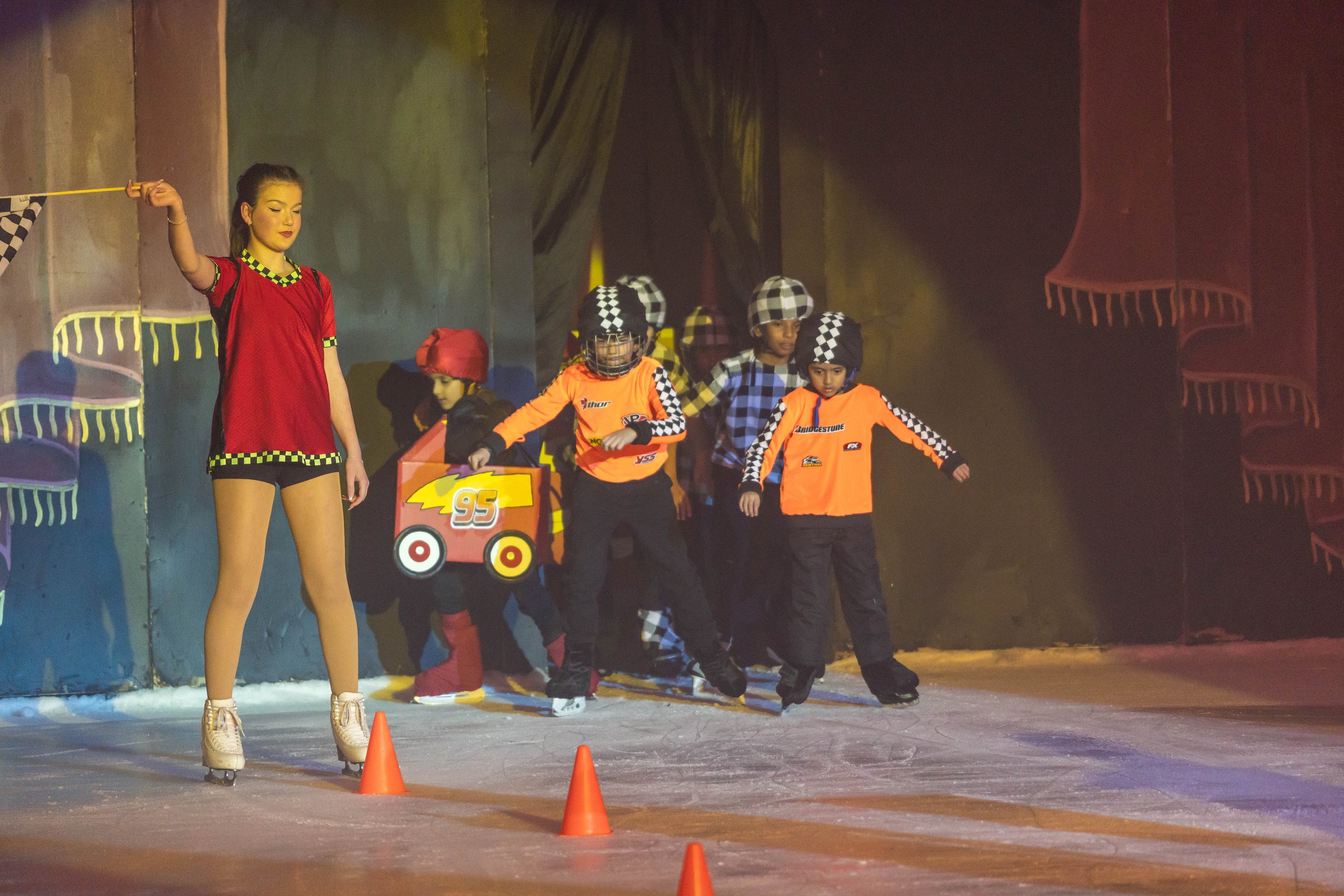 A group of children dressed as race car drivers participating in an ice skating race, guided by a woman in a black and white checkered outfit, on an indoor ice rink.