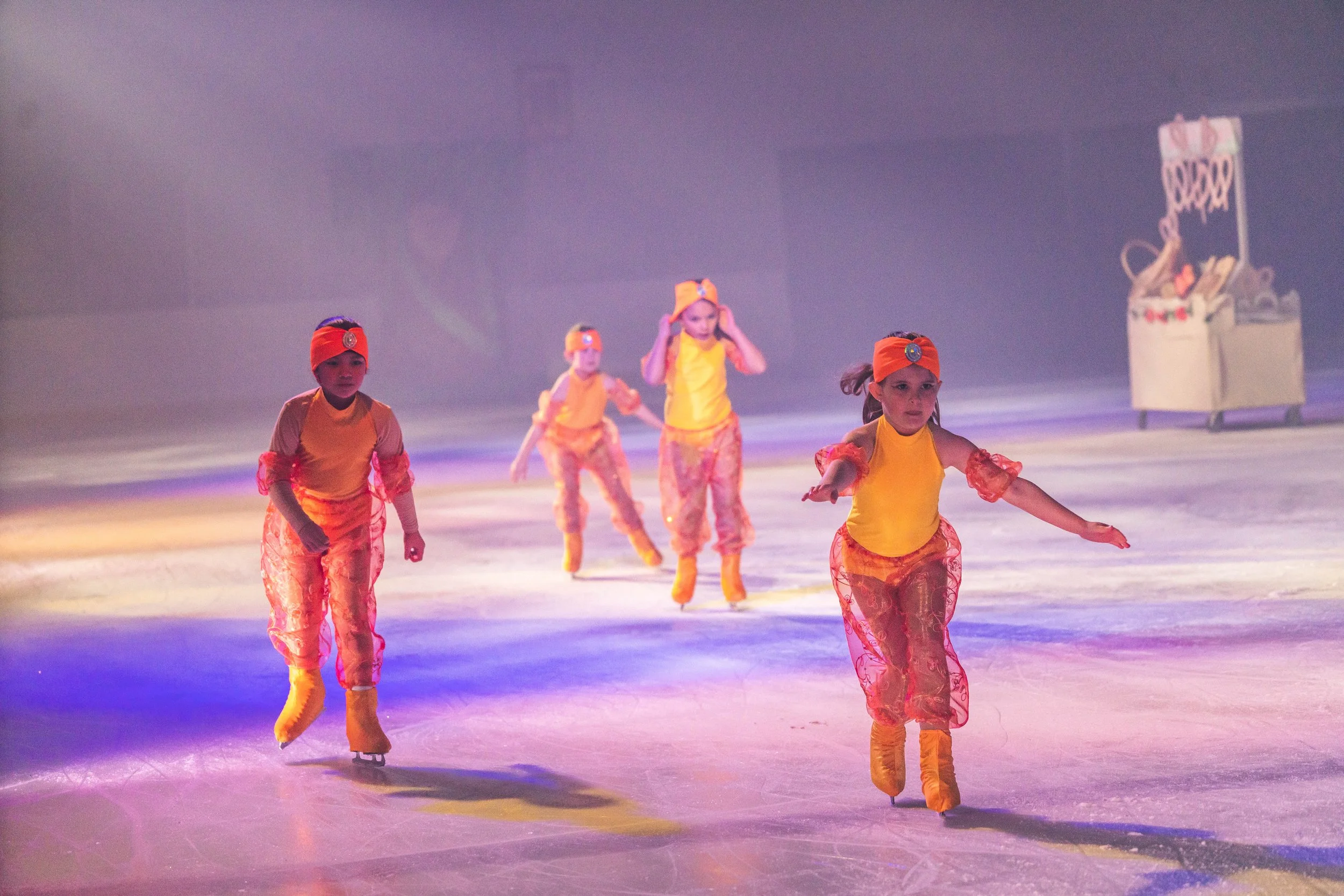 Young children dressed in colorful costumes ice skating on an indoor rink with a purple background and a decorative prop in the background.