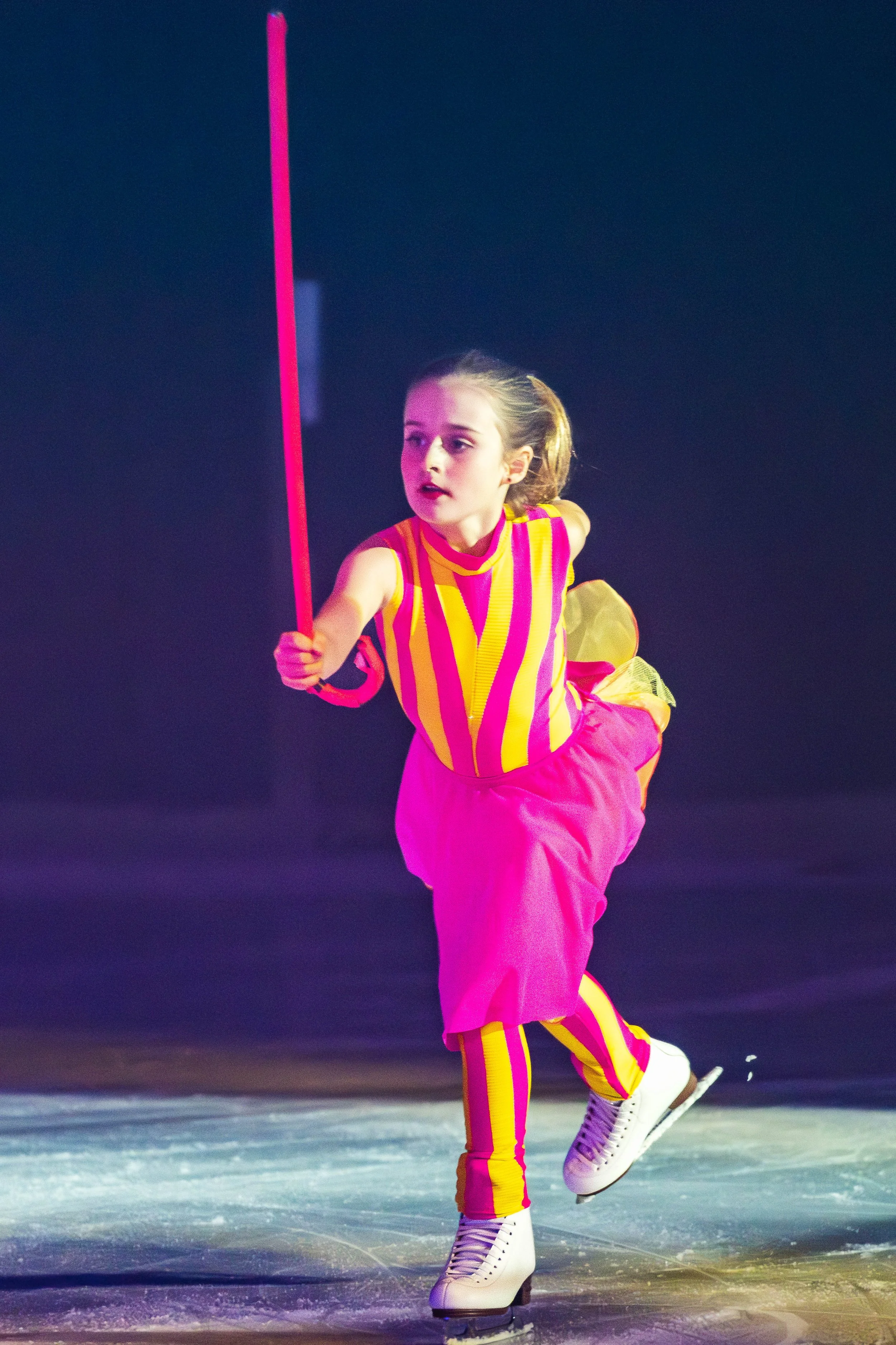 A young girl ice skating on an ice rink, holding a pink stick, dressed in a colorful striped top and bright pink pants, wearing white ice skates.