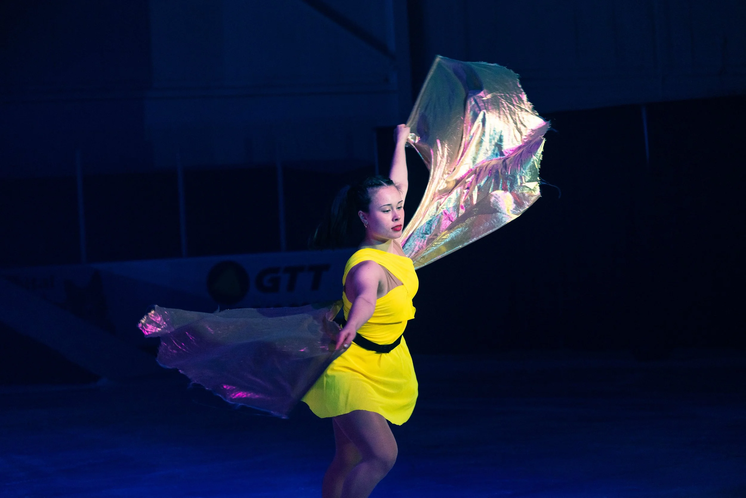 A young woman in a yellow dress performing a dance with iridescent fabric in a dark, indoor setting.