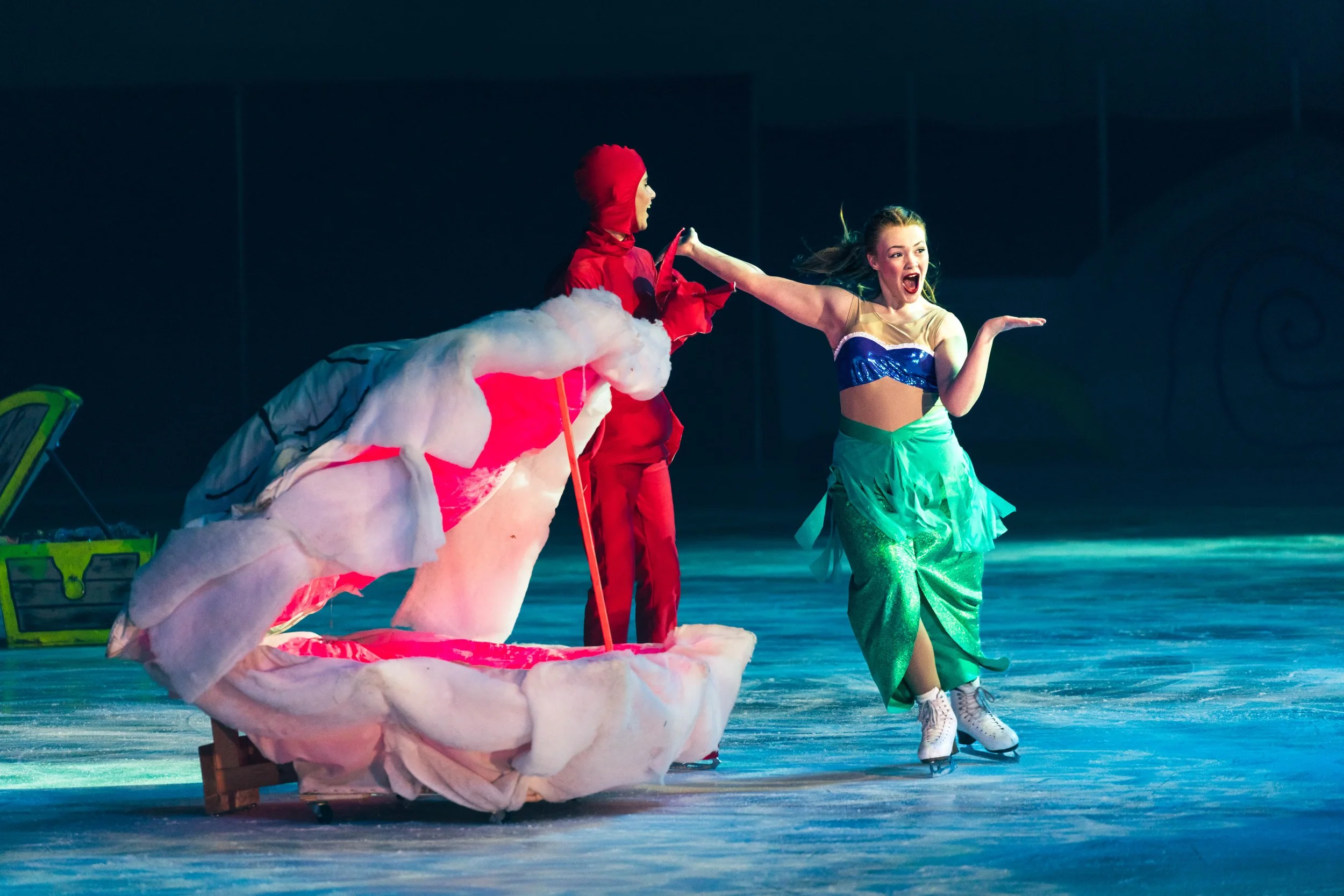 A performer dressed as a colorful, whimsical character with a pink and white costume and a red hood icicles, ice skates, appears to be on an ice rink. Another performer in a bright, shiny costume with a green skirt and a blue top is ice skating, laug