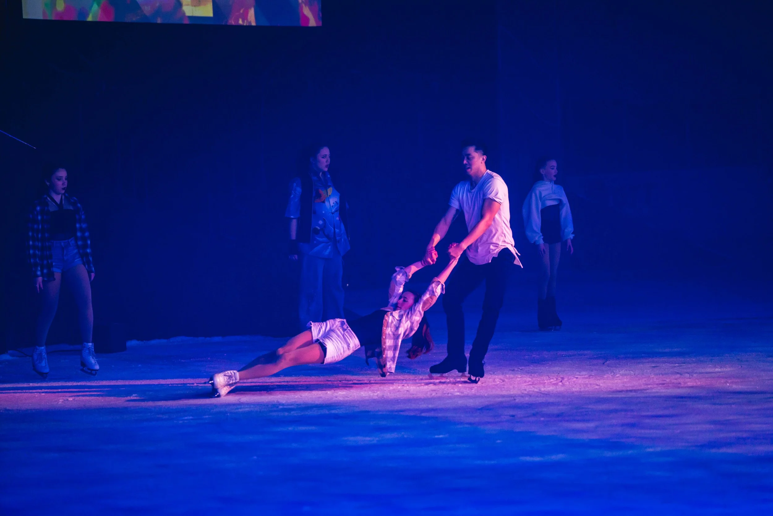 A scene from a theatrical performance with five performers under blue lighting. Two performers stand, and one performer is on the ground, holding hands with a standing performer. The scene suggests a dance or drama act.