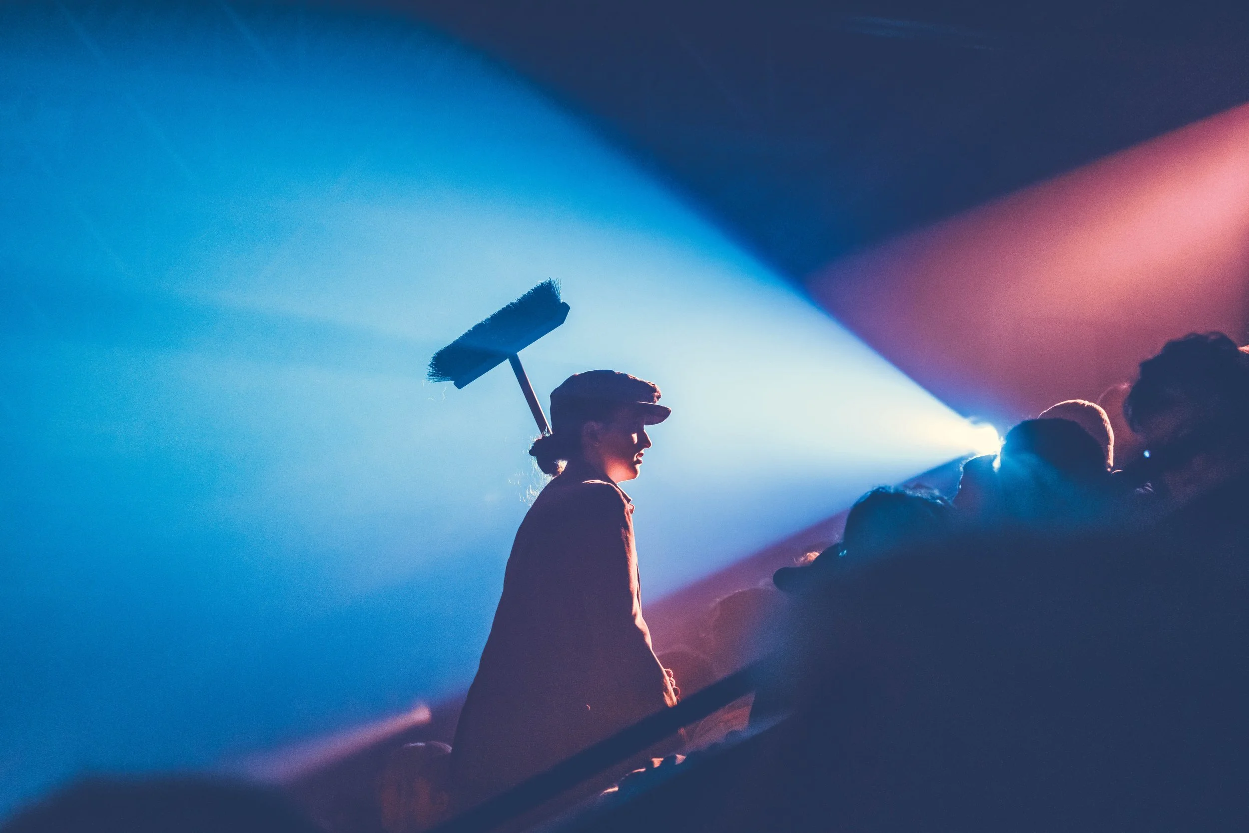 Side profile of a woman wearing a baseball cap, holding a broom, standing in a crowd with vibrant stage lighting.