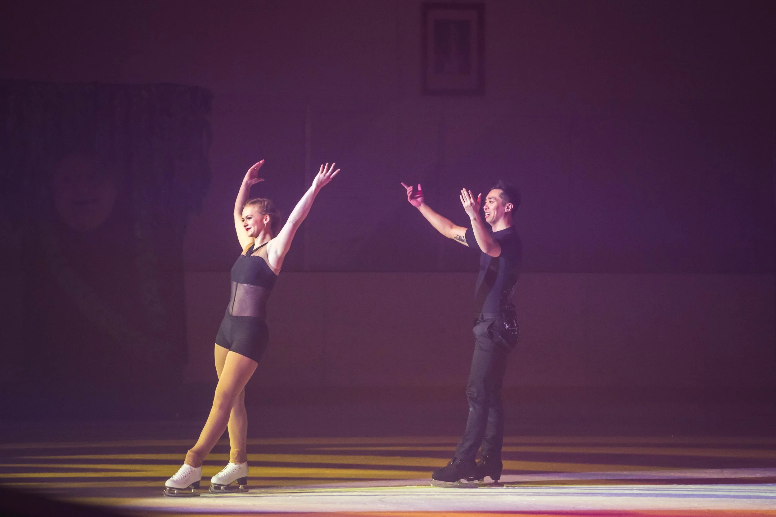 A man and a woman figure skating indoors under purple lighting, with the man standing behind the woman and both raising their arms in a pose.