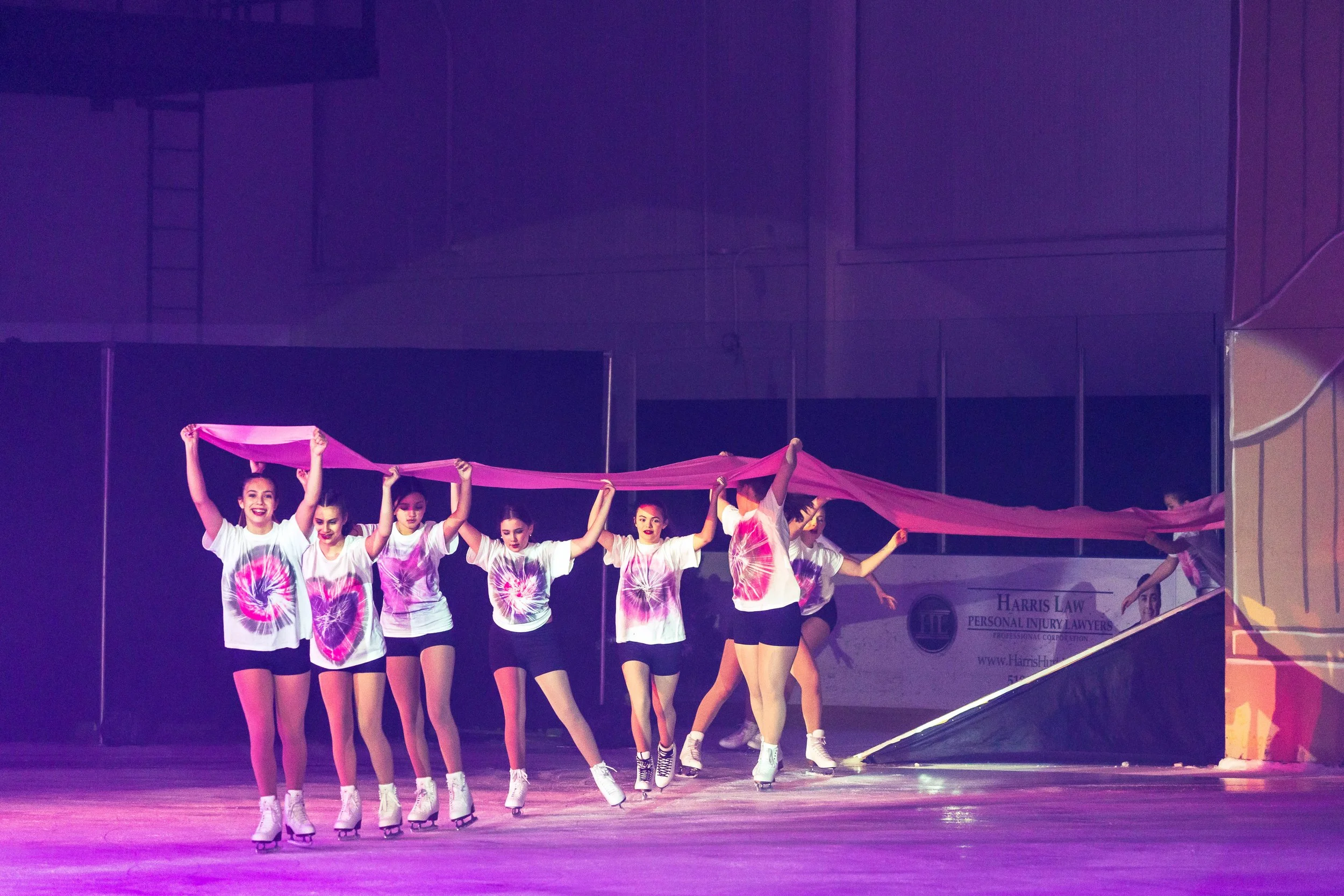 A group of young women performing a dance on stage, wearing matching tie-dye t-shirts and black shorts, holding a long pink fabric above their heads, with stage lighting illuminating them.