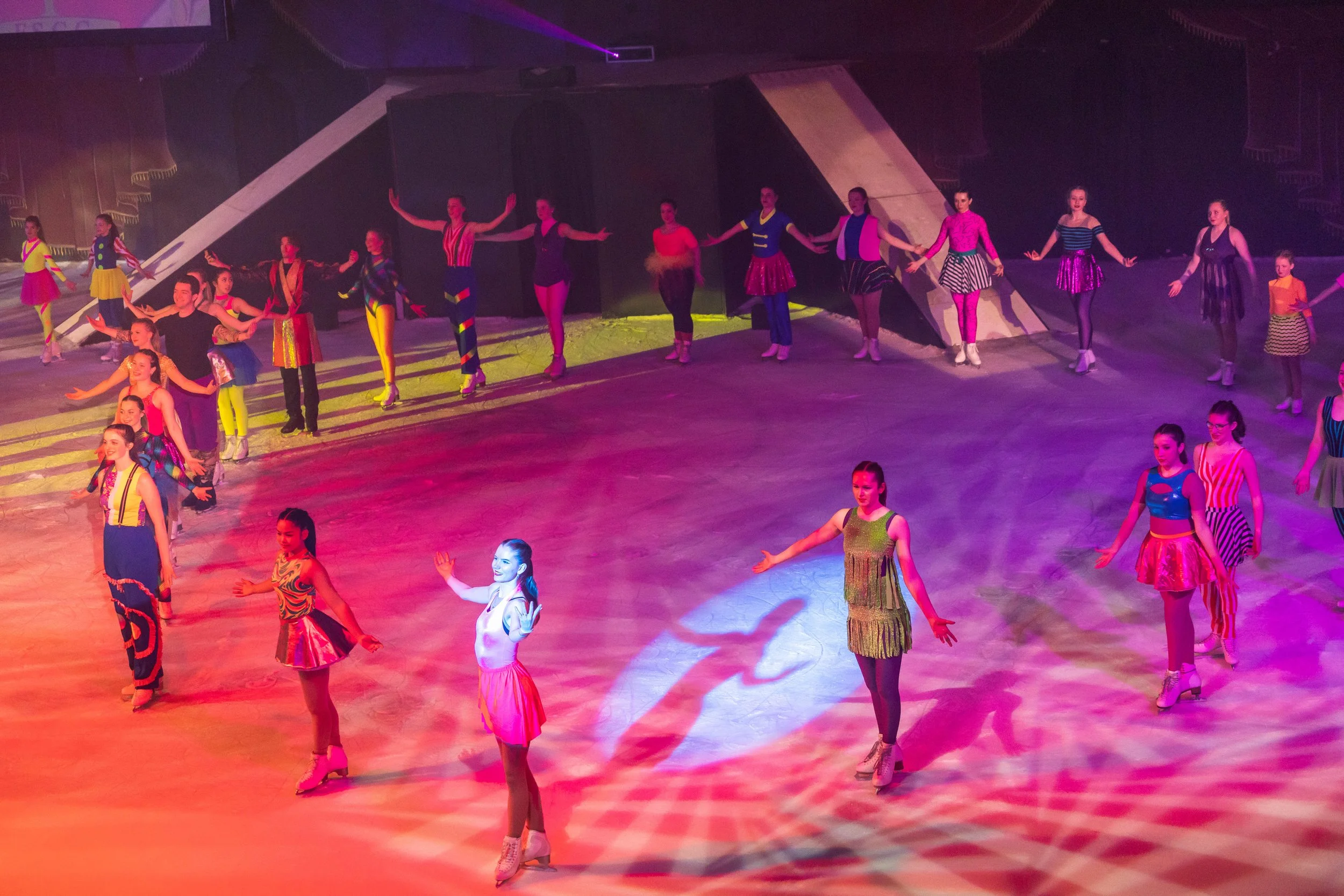 A group of performers, mostly women, dressed in colorful costumes, holding hands and standing in a line on stage with colored lights and a dark background.