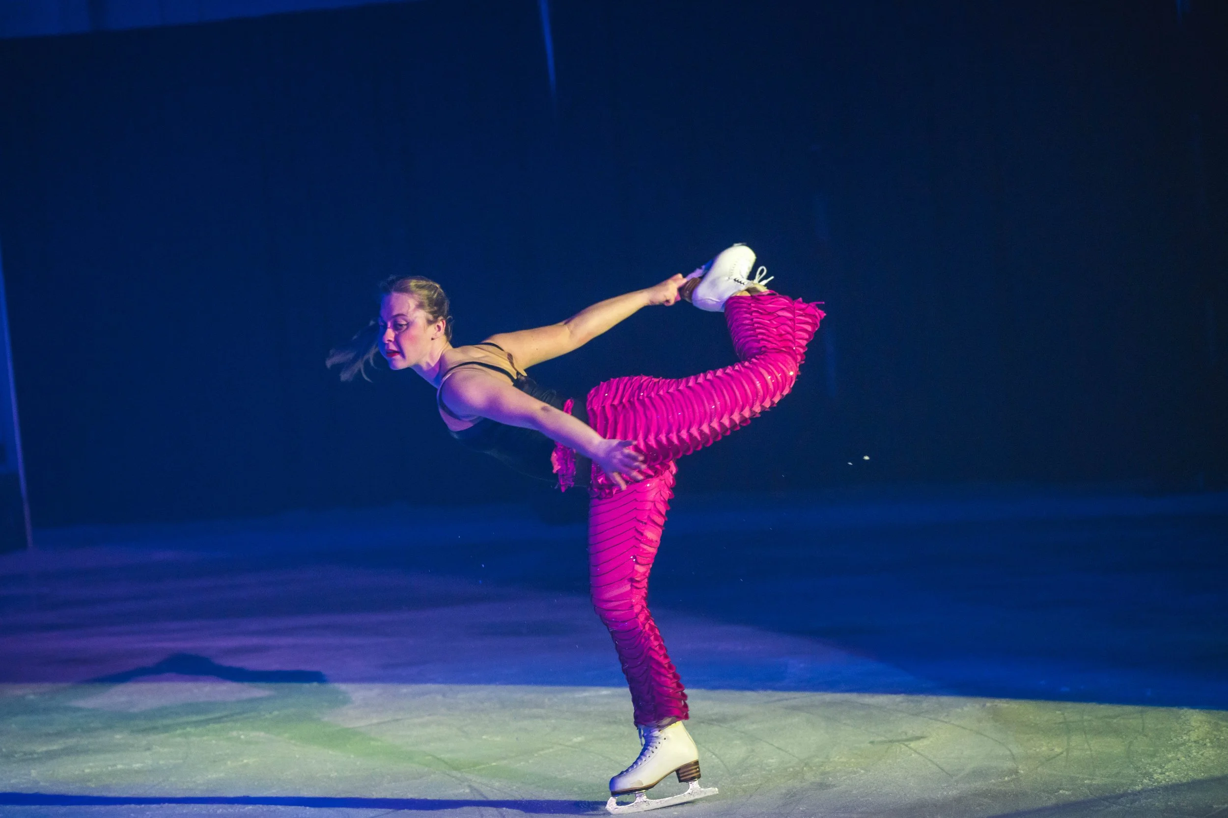 A female figure skater performing a difficult move on the ice, balancing on one leg while holding the blade of her skate with her hand, dressed in bright pink pants and a tan top, with a dark background and colorful lighting on the ice.