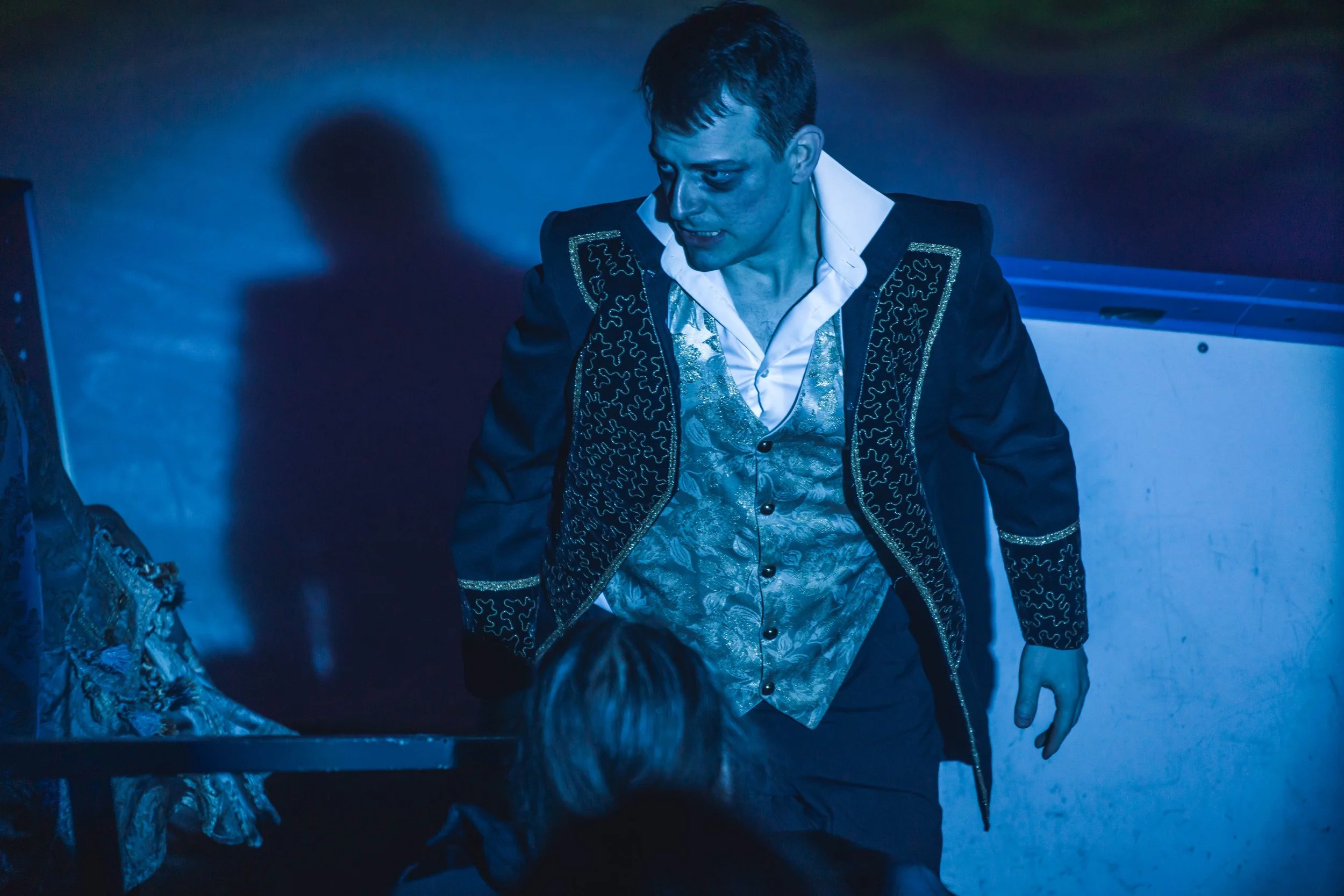 A man wearing a detailed suit with gold and silver embroidery, illuminated by blue lighting, leaning over a surface with his right hand resting on it.