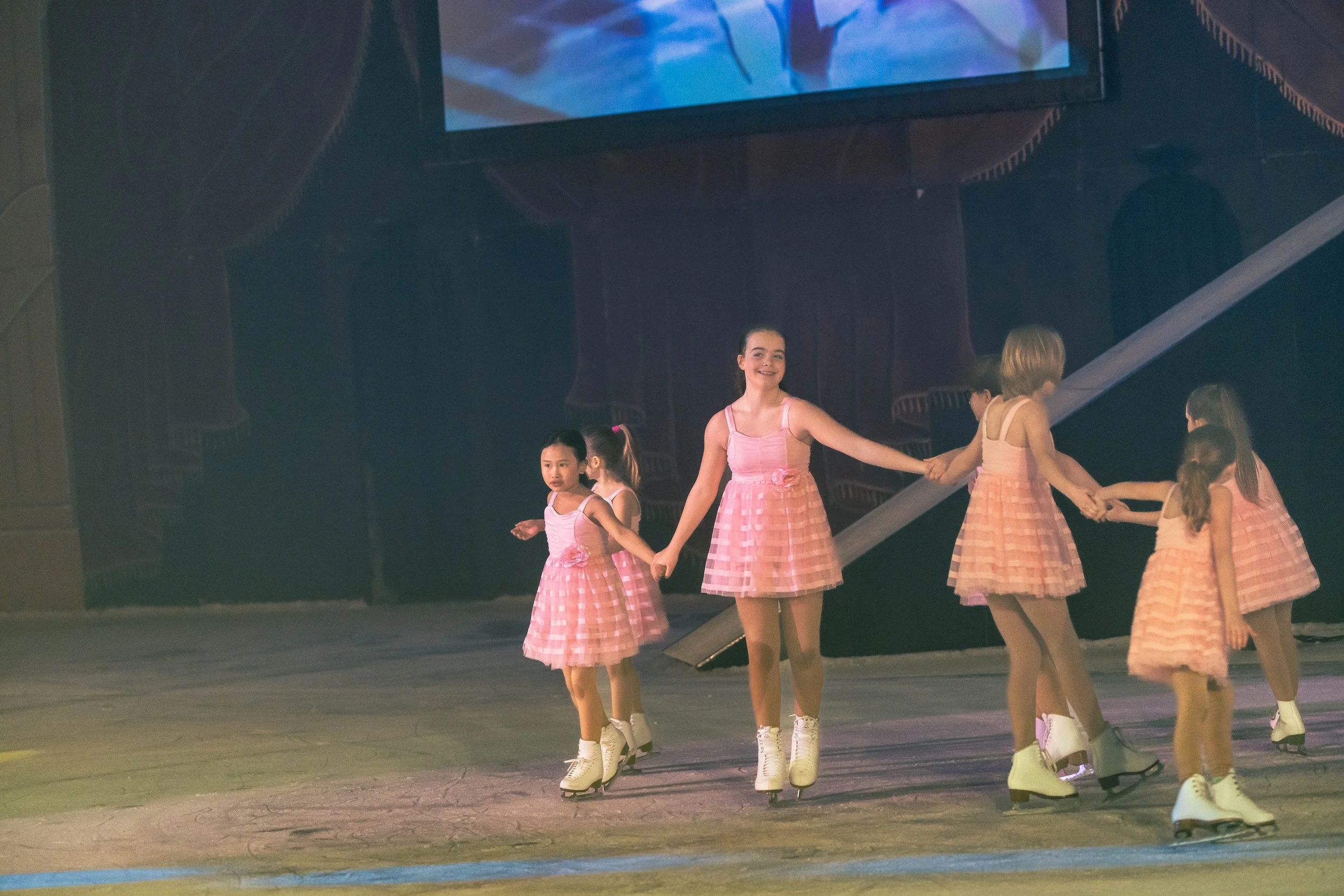 A group of young girls ice skating in pink dresses holds hands on an indoor ice rink.