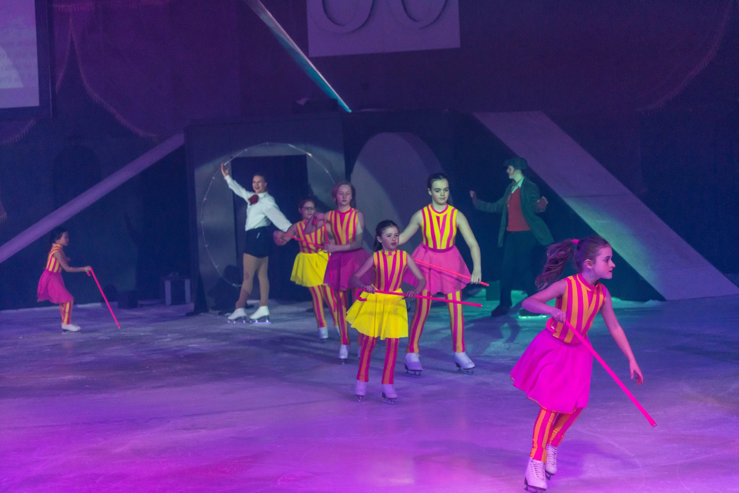 Group of young girls dressed in colorful striped costumes performing roller skating dance routine on stage, with a man and woman in the background.
