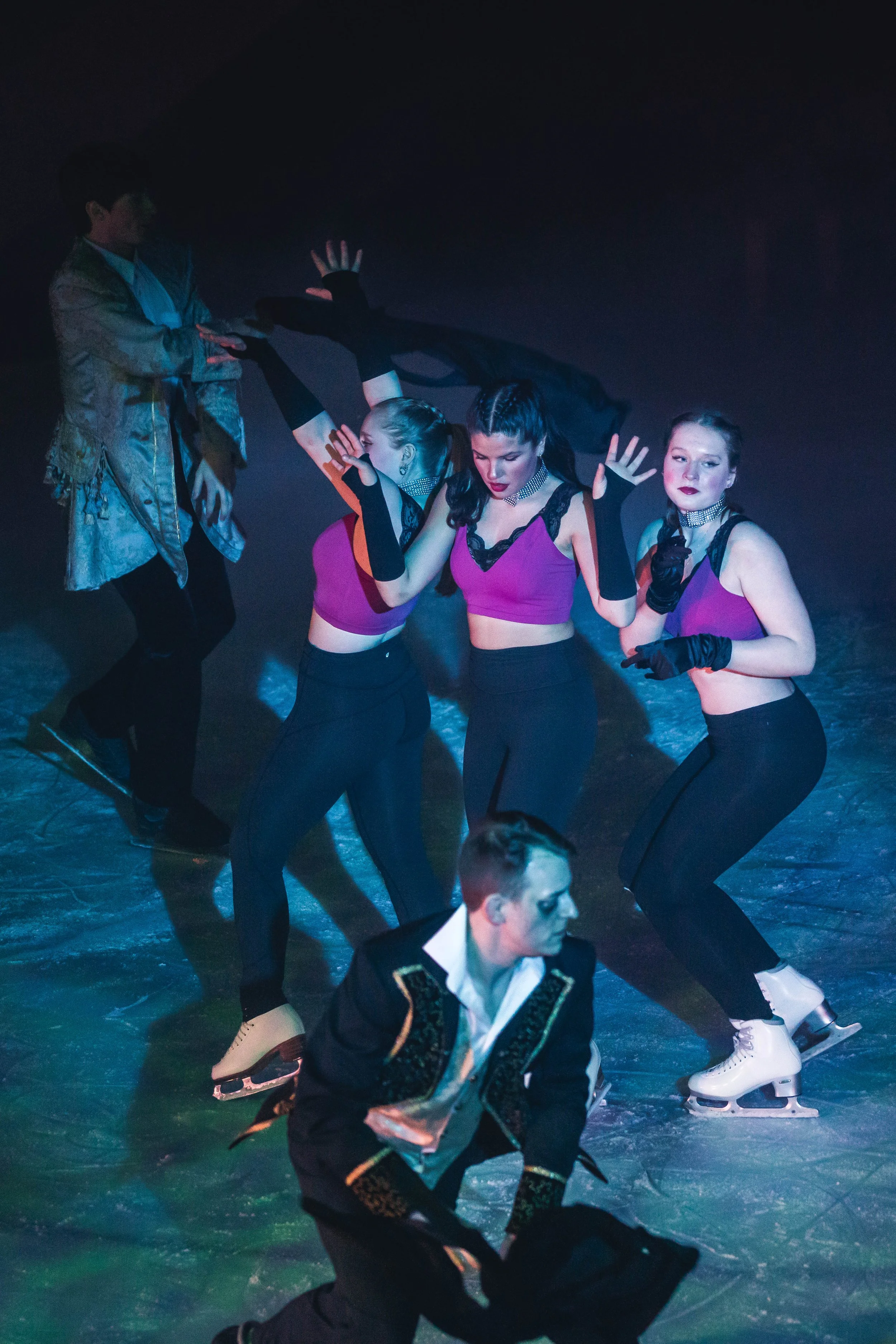 Four ice skaters practicing figure skating moves in a dark setting, two women in purple tops with black pants and two men, one in a detailed black jacket and another in a shiny shirt.