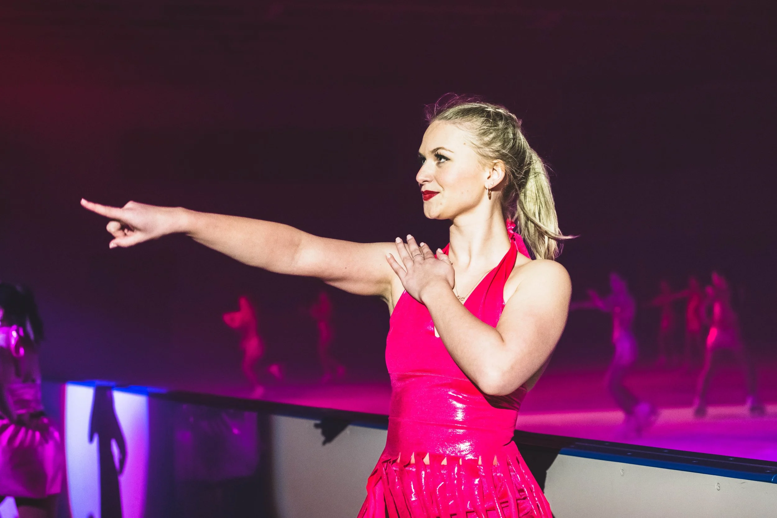 A woman in a bright pink dress performing a dance move with her right arm extended and her left hand on her chest, inside a dimly lit venue with purple and pink lighting.