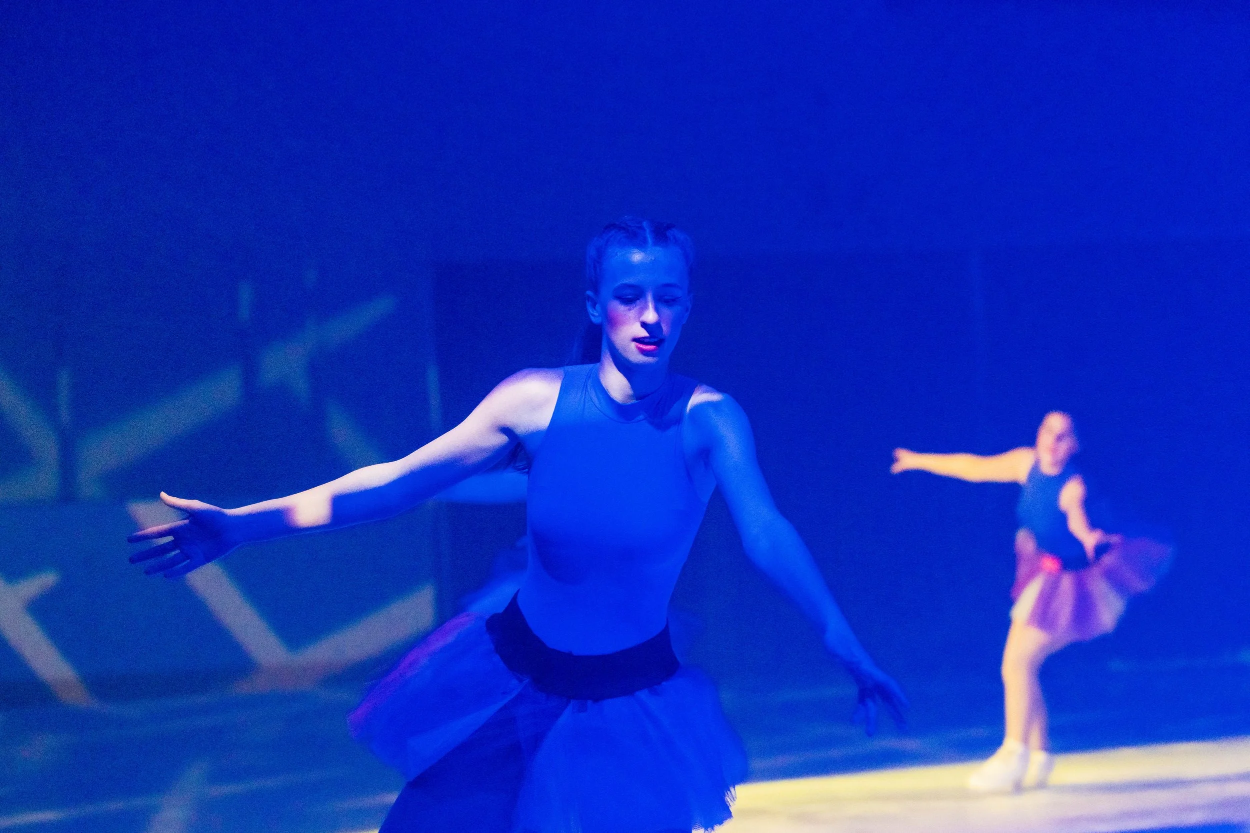 A ballerina practicing on stage under blue lighting, with arms extended and wearing a black leotard and tutu.