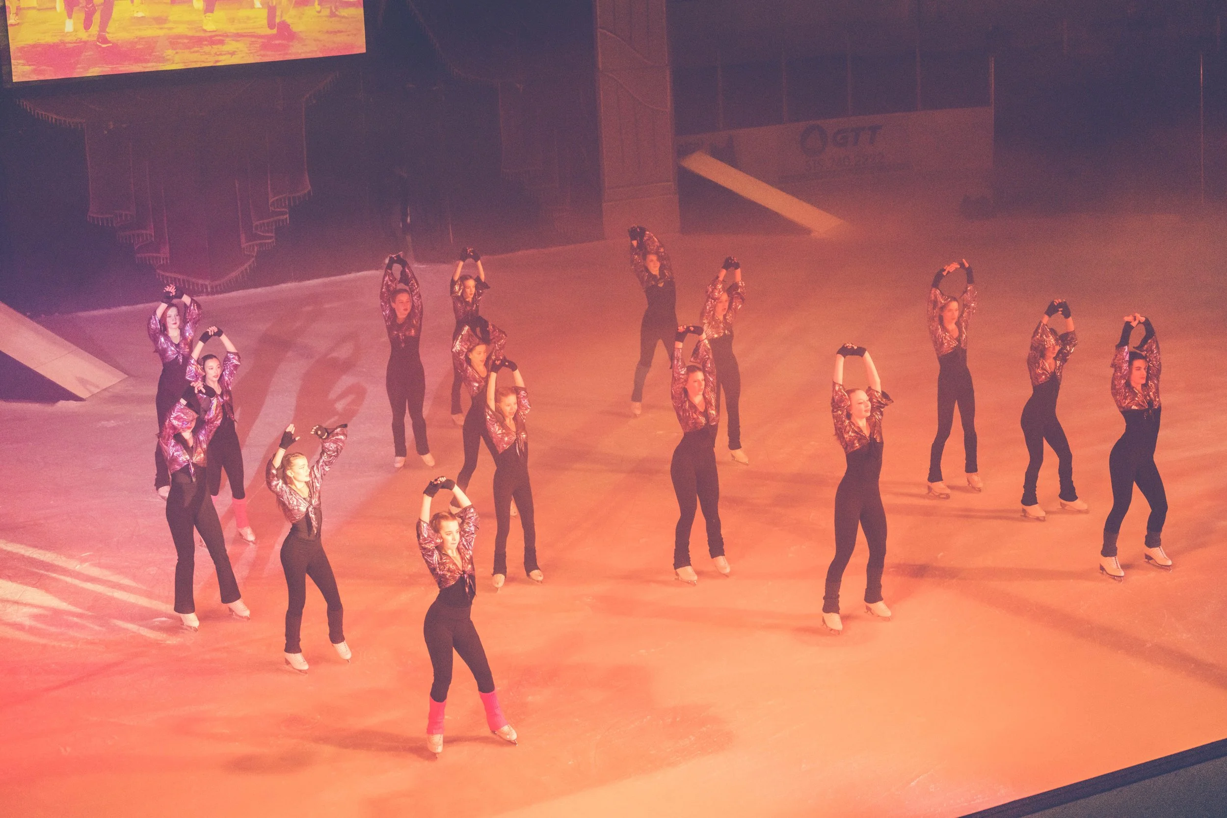 A group of performers wearing black pants and shiny, patterned tops on skates, posing and stretching on an ice rink during a performance or practice. There is a large screen above and behind them showing colorful graphics.