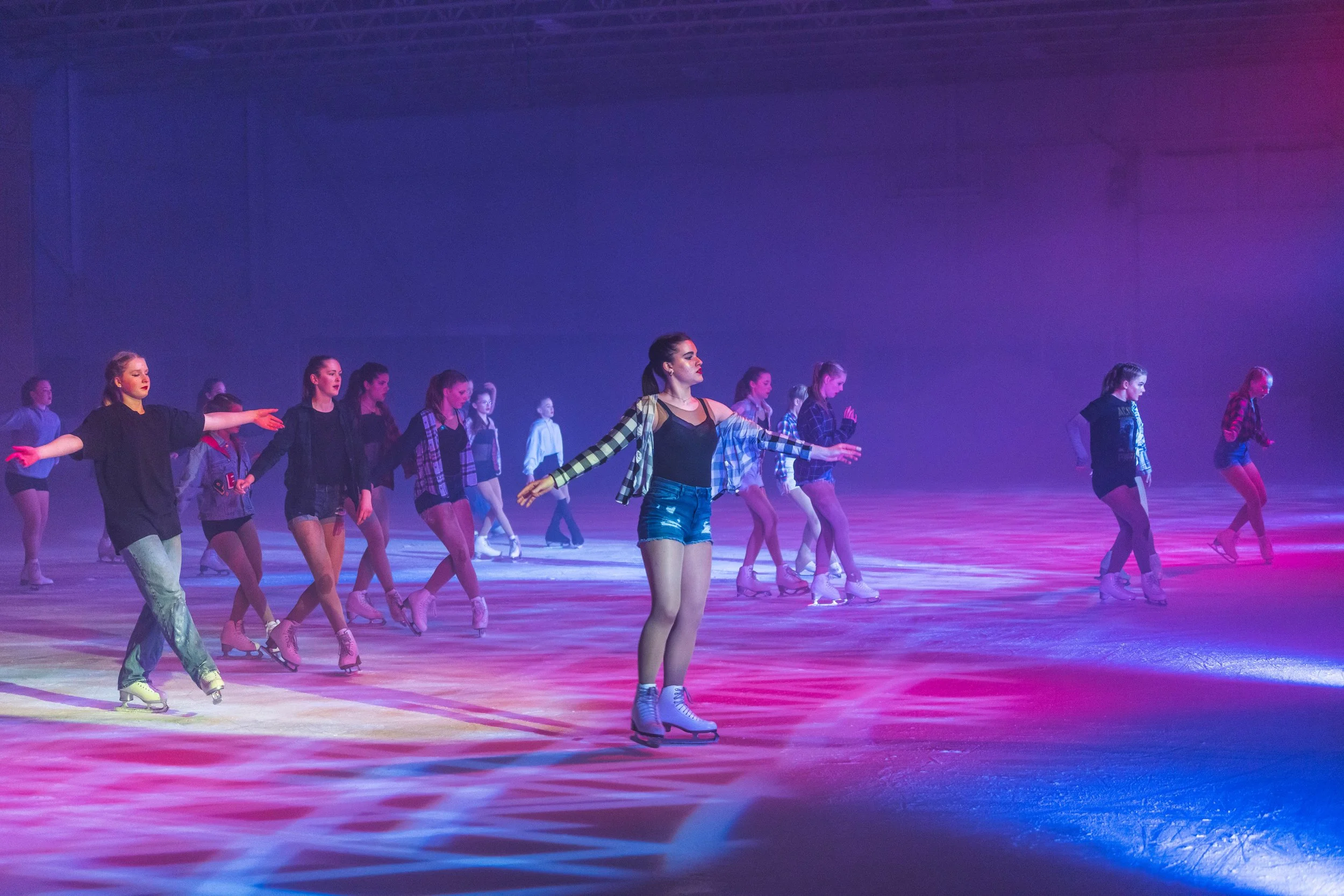 A group of young women figure skating on an indoor rink under colorful stage lighting.