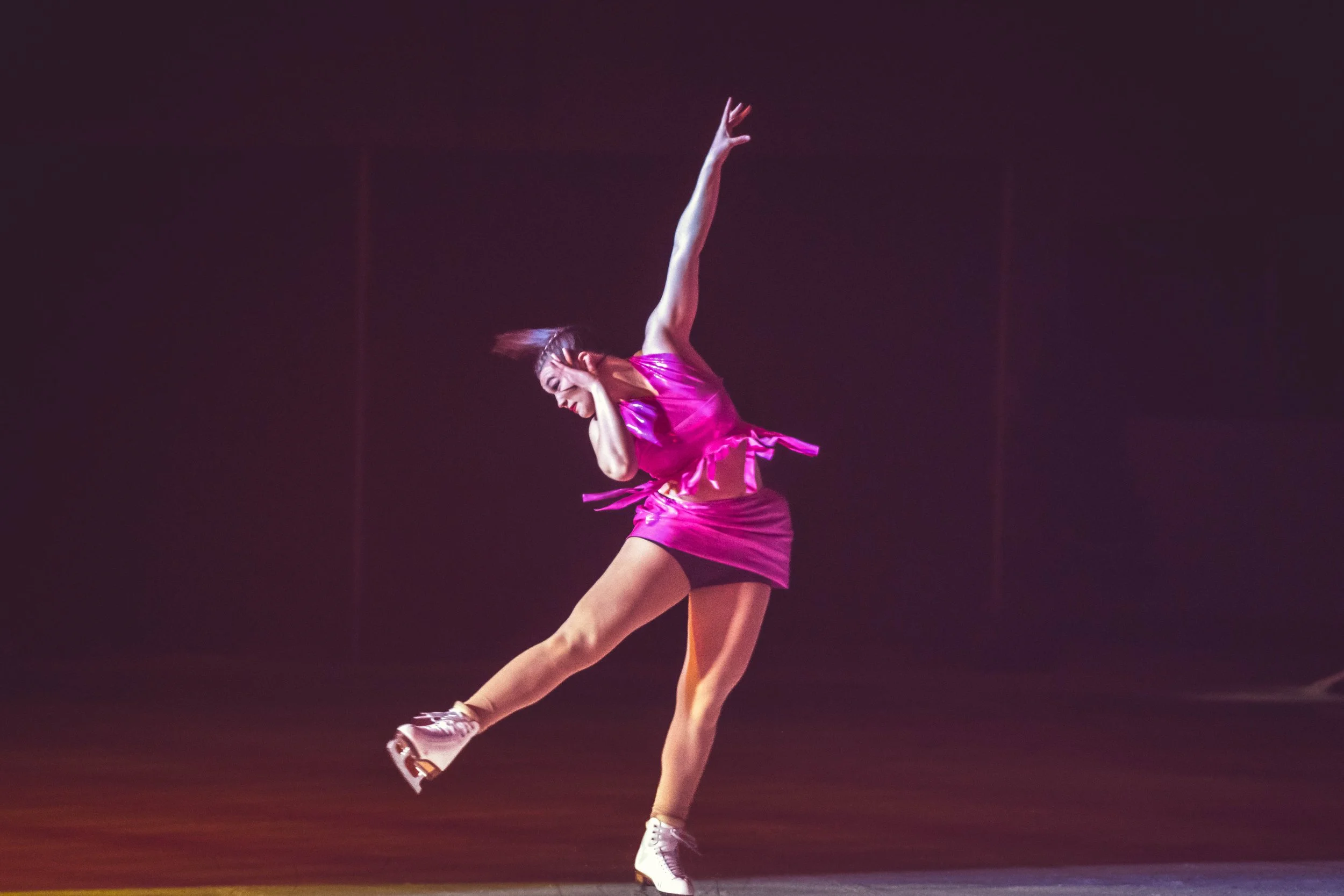 A female dancer in a pink dress is performing a dance move, balancing on her left foot with her right leg extended outward. She is holding her head with her left hand and has her right arm raised above her head. The background is dark with stage ligh