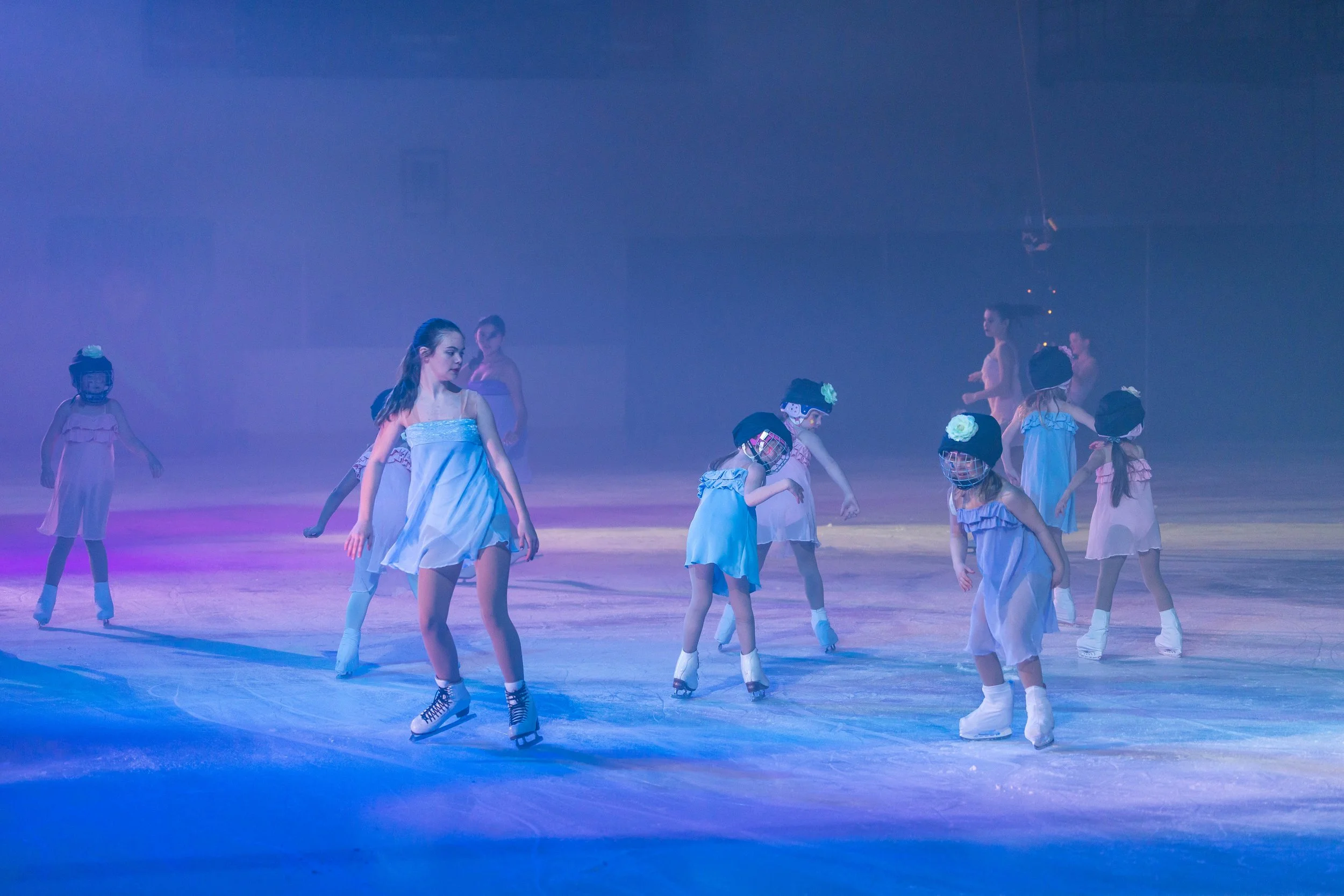 Children wearing costumes and skates practicing ice skating in a rink under colored lighting.