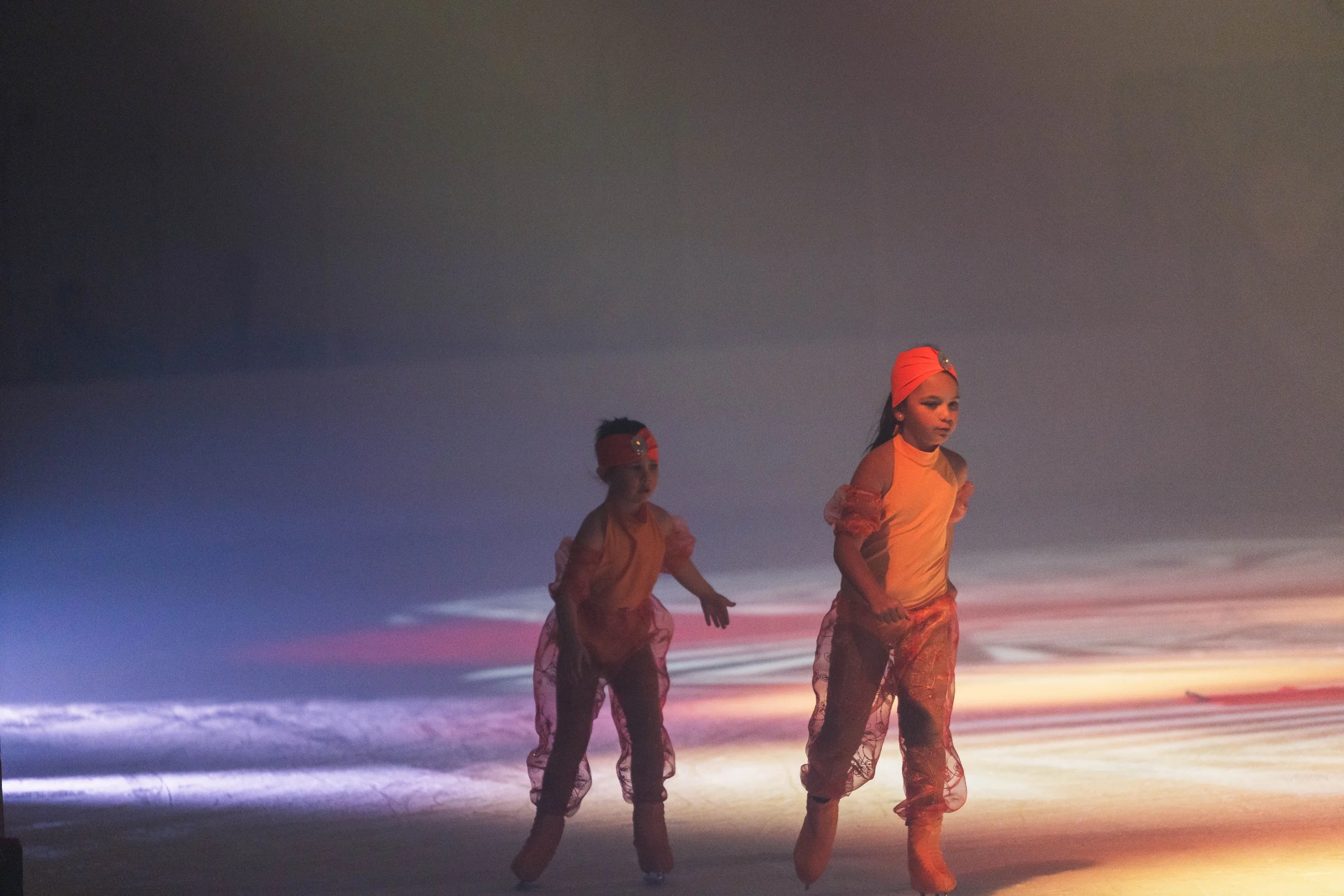 Two dancers wearing orange costumes and orange head wraps perform on a beach at sunset, illuminated by colorful lighting.