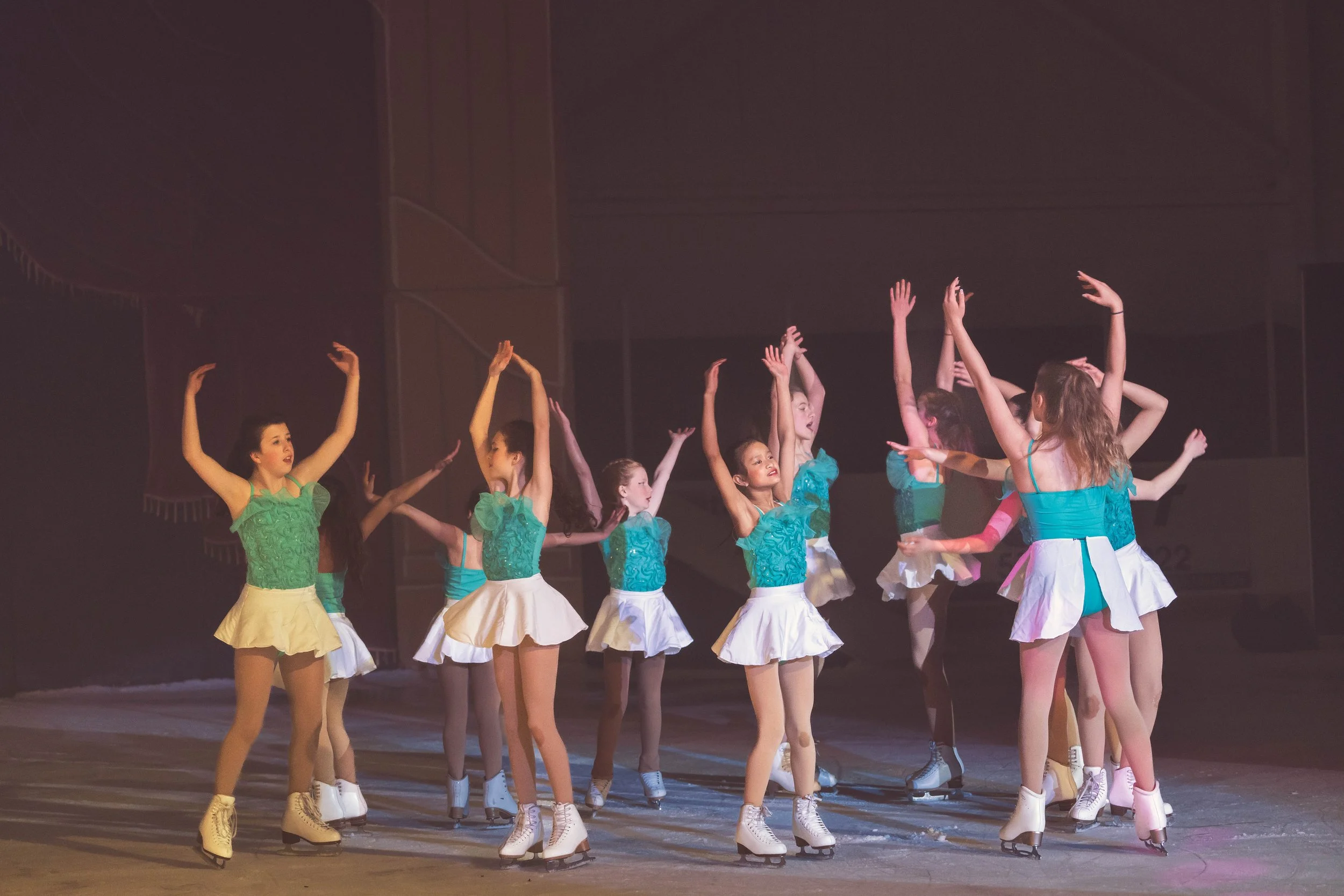 Group of young female figure skaters in colorful costumes practicing ballet moves on ice.