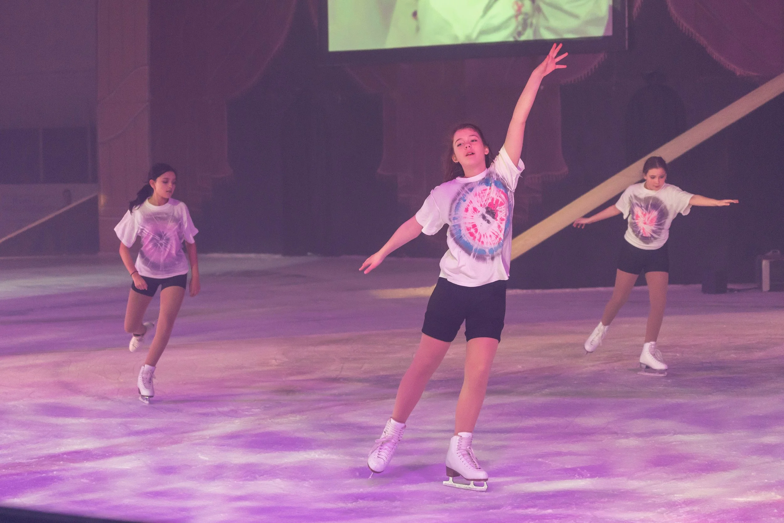 Three girls ice skating on an indoor rink, one in the center with her arm raised and wearing a tie-dye t-shirt, the other two on either side with their arms extended, all wearing ice skates.