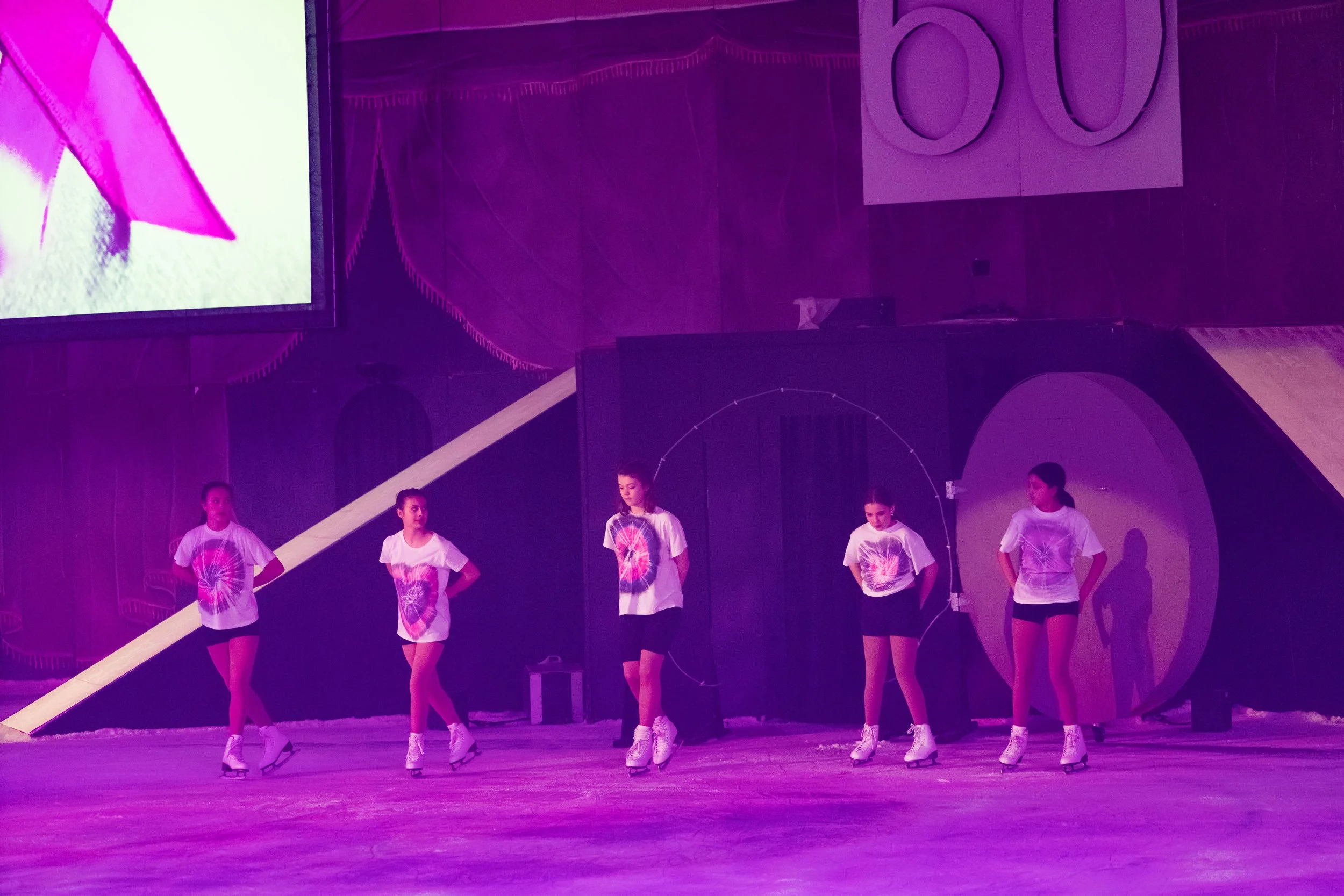 A group of young girls wearing matching white t-shirts with colorful tie-dye designs, white ice skates, and black shorts, standing in a line on an indoor roller skating rink under purple lighting.