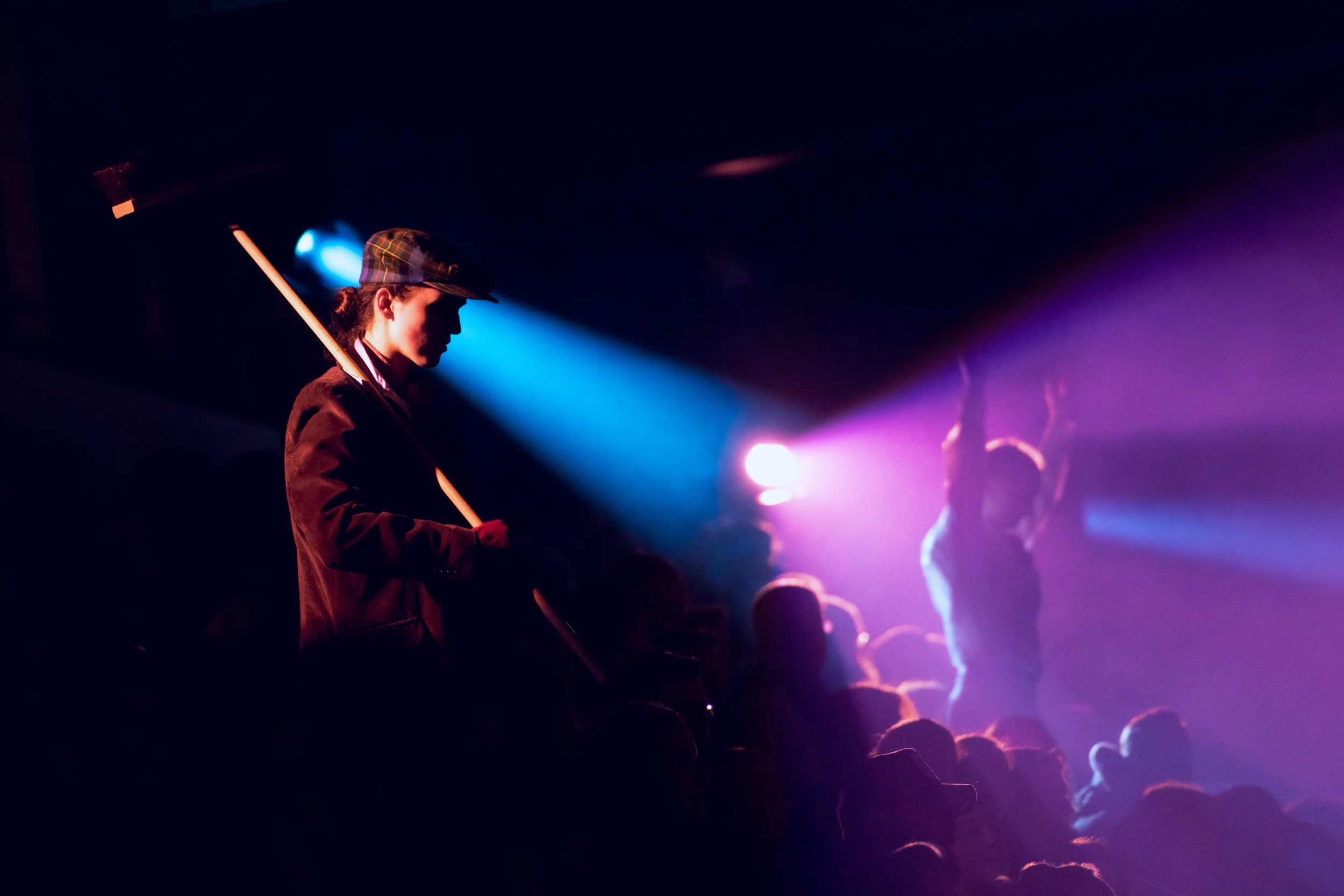 A person in a hat and jacket holding a broomstick, standing silhouetted against colorful stage lighting at a concert or event.