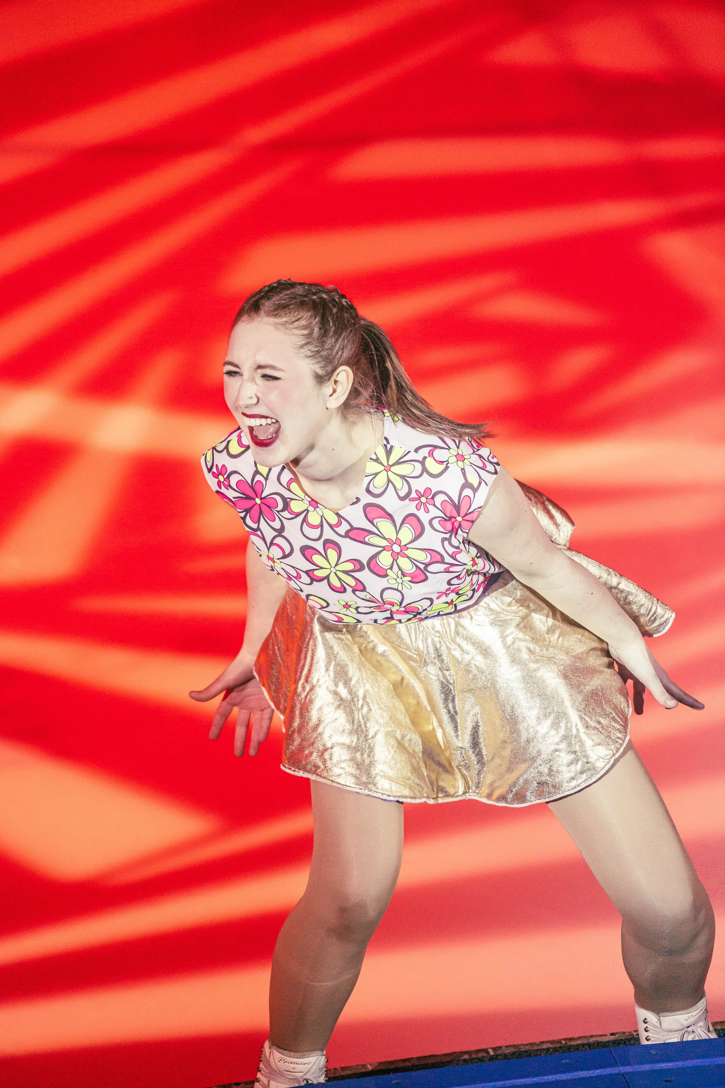 A young woman in a colorful floral top and shiny gold skirt is performing an energetic dance move with a vibrant orange background.