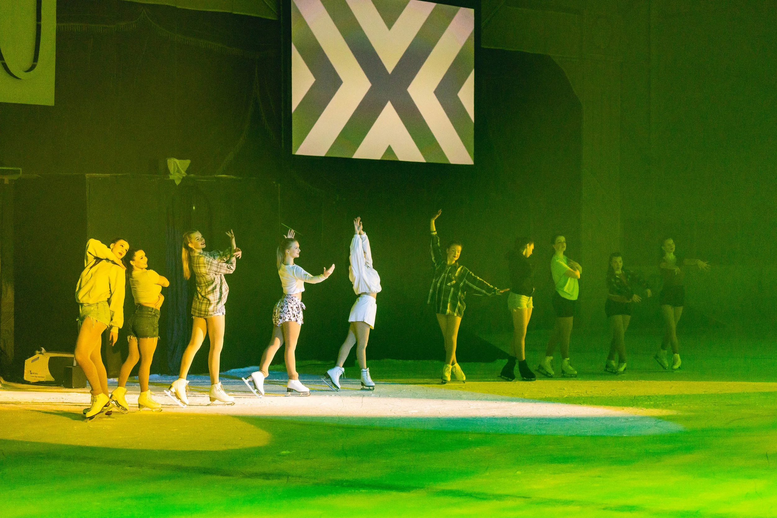 A group of people in colorful casual attire wearing ice skates, standing in a line on an ice rink, with a large geometric light projection on the wall behind them, illuminated with yellow and green lighting.
