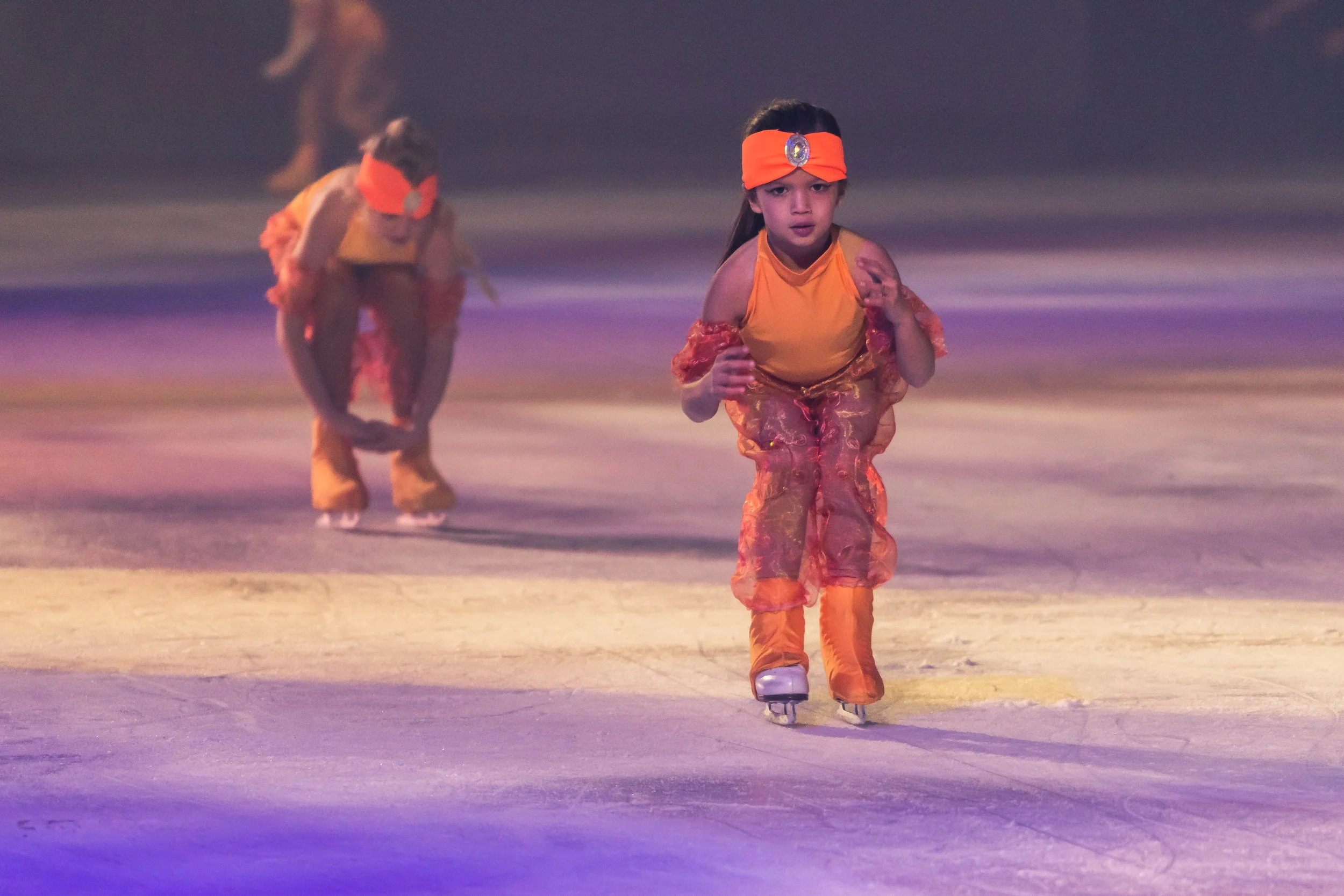 Two children ice skating, dressed in orange costumes with headbands, on an ice rink with colorful lighting.