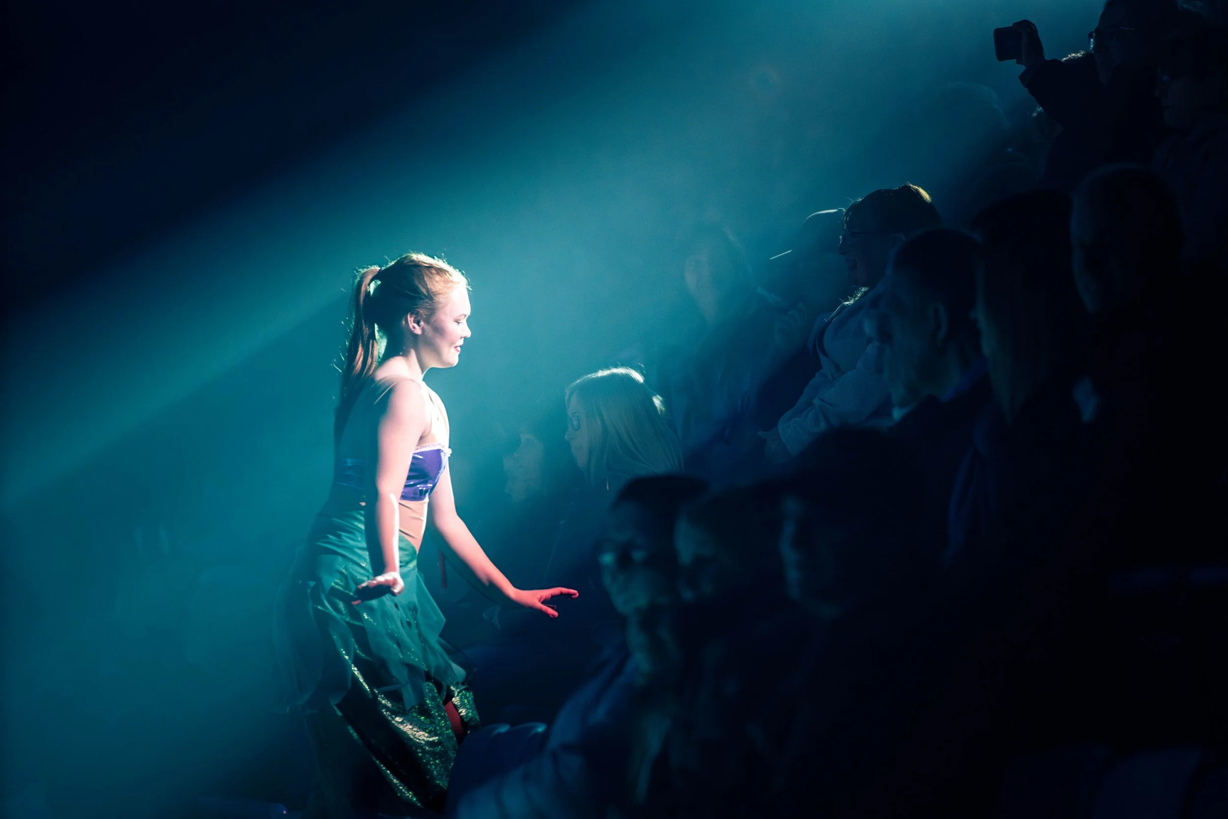 A young girl standing on a stage with her eyes closed and a serene expression, illuminated by a spotlight, surrounded by an audience in darkness.