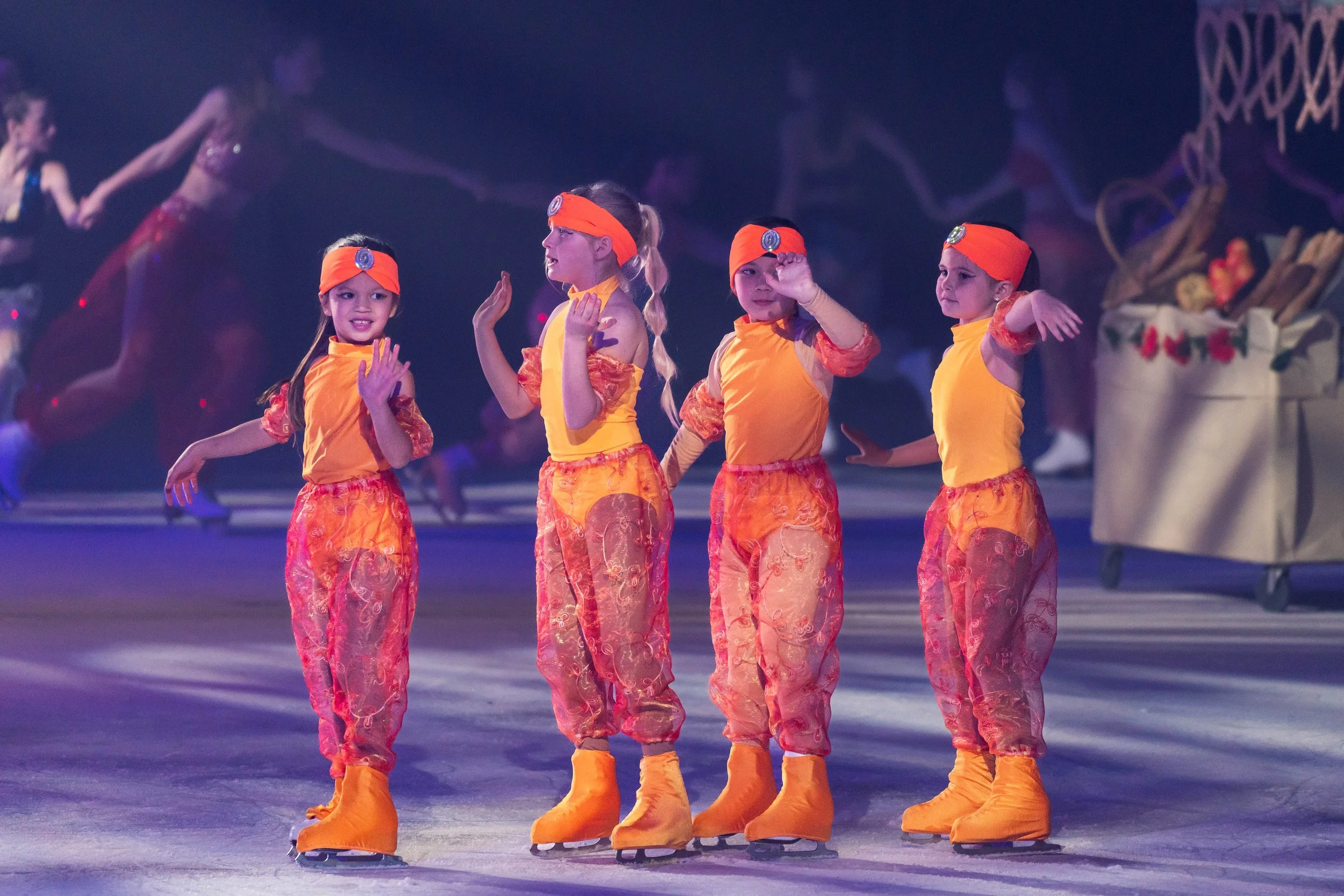 Four young girls in matching orange and yellow costumes, standing on ice skates in a line, holding hands, with a decorative holiday-themed backdrop in the background.