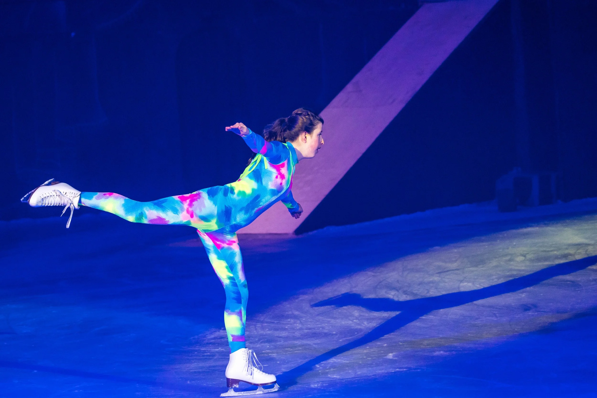 A woman figure skater in a colorful tie-dye outfit performs an arabesque on ice, with one leg extended behind her and arms outstretched.