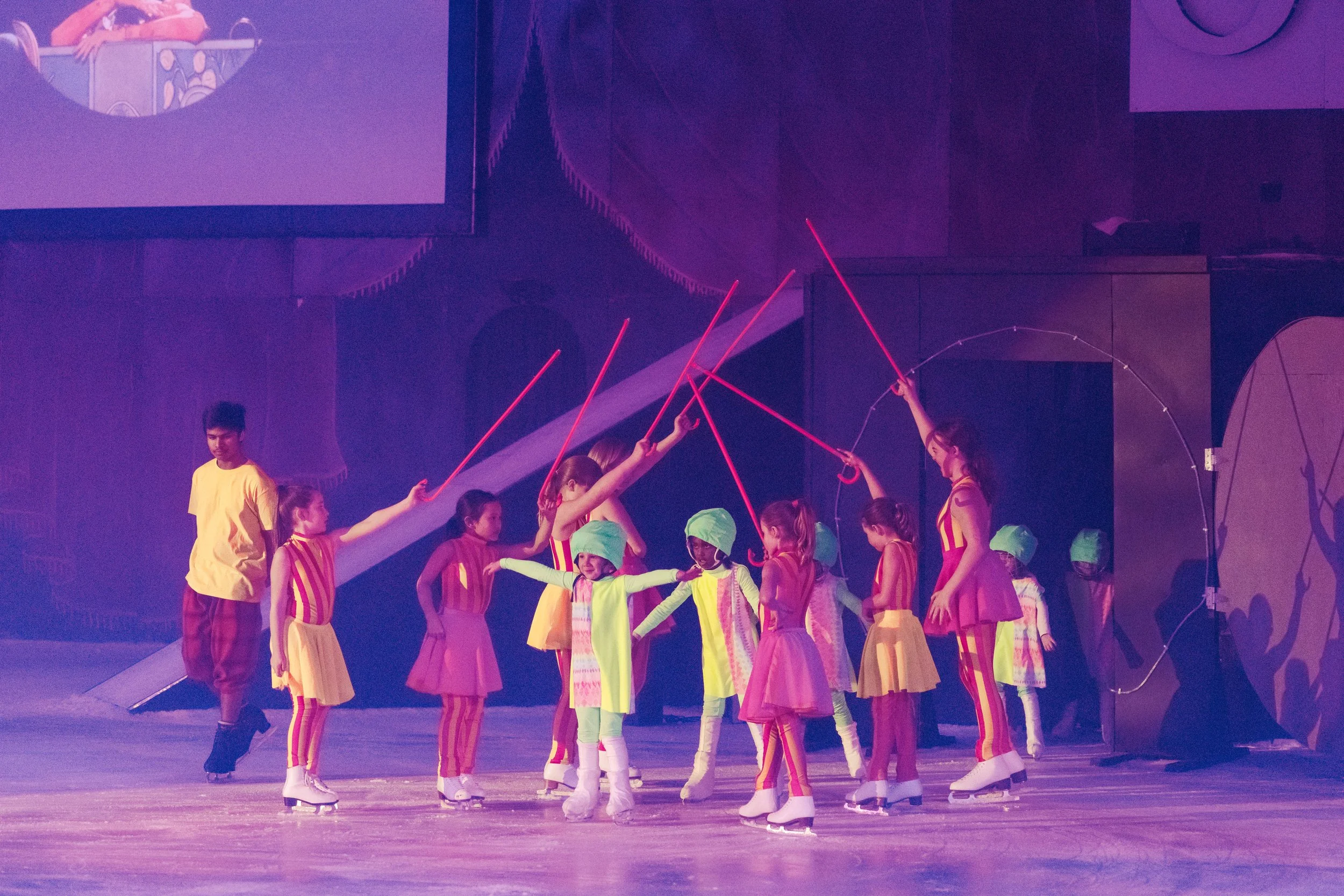 Children wearing colorful costumes and ice skates performing a routine on an ice rink during a show. Some children are holding pink glow sticks while a few adults are assisting or observing.