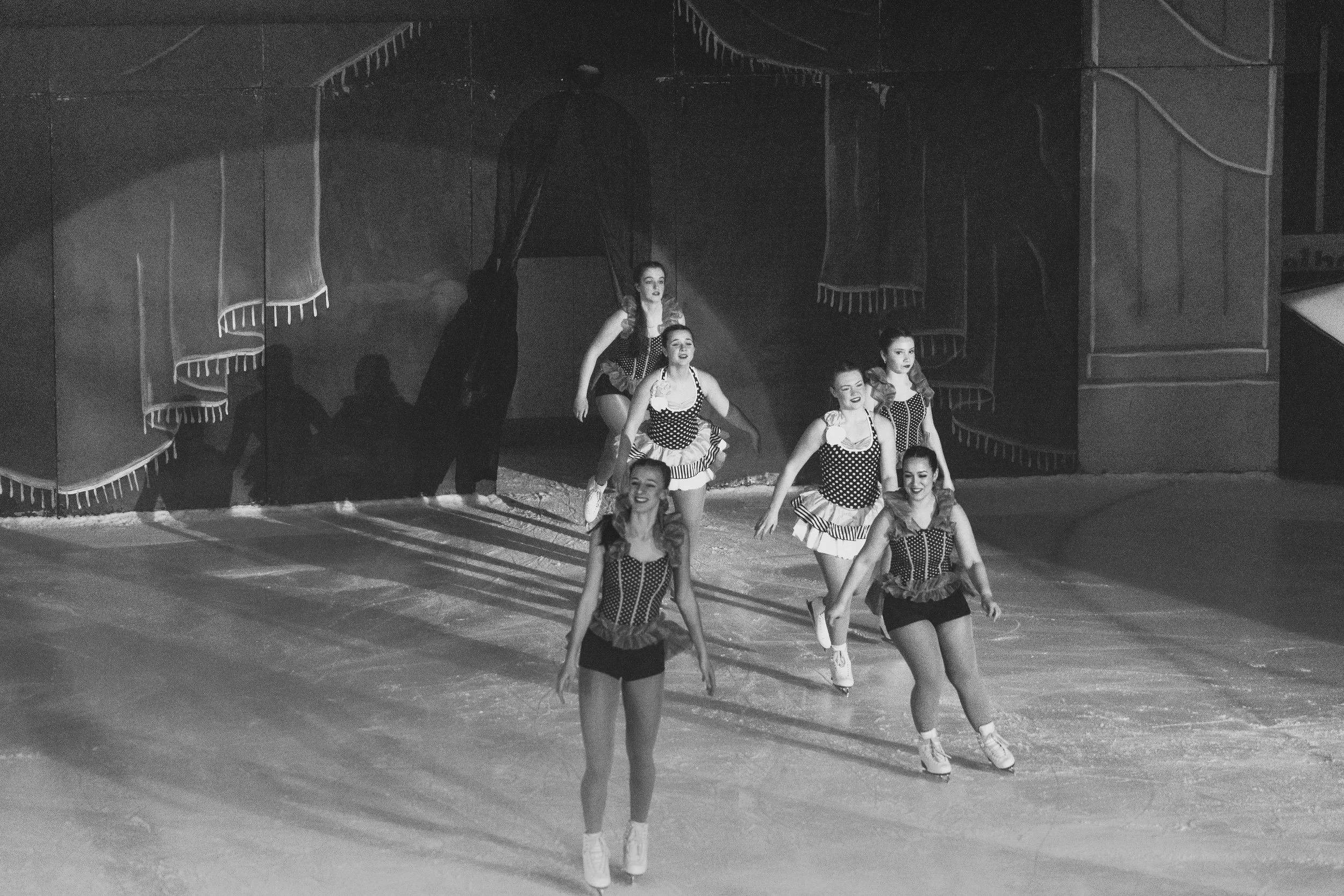 Black and white photo of a group of female performers in vintage-style costumes, standing and dancing in formation on stage, with a stage backdrop that suggests a circus or carnival theme.