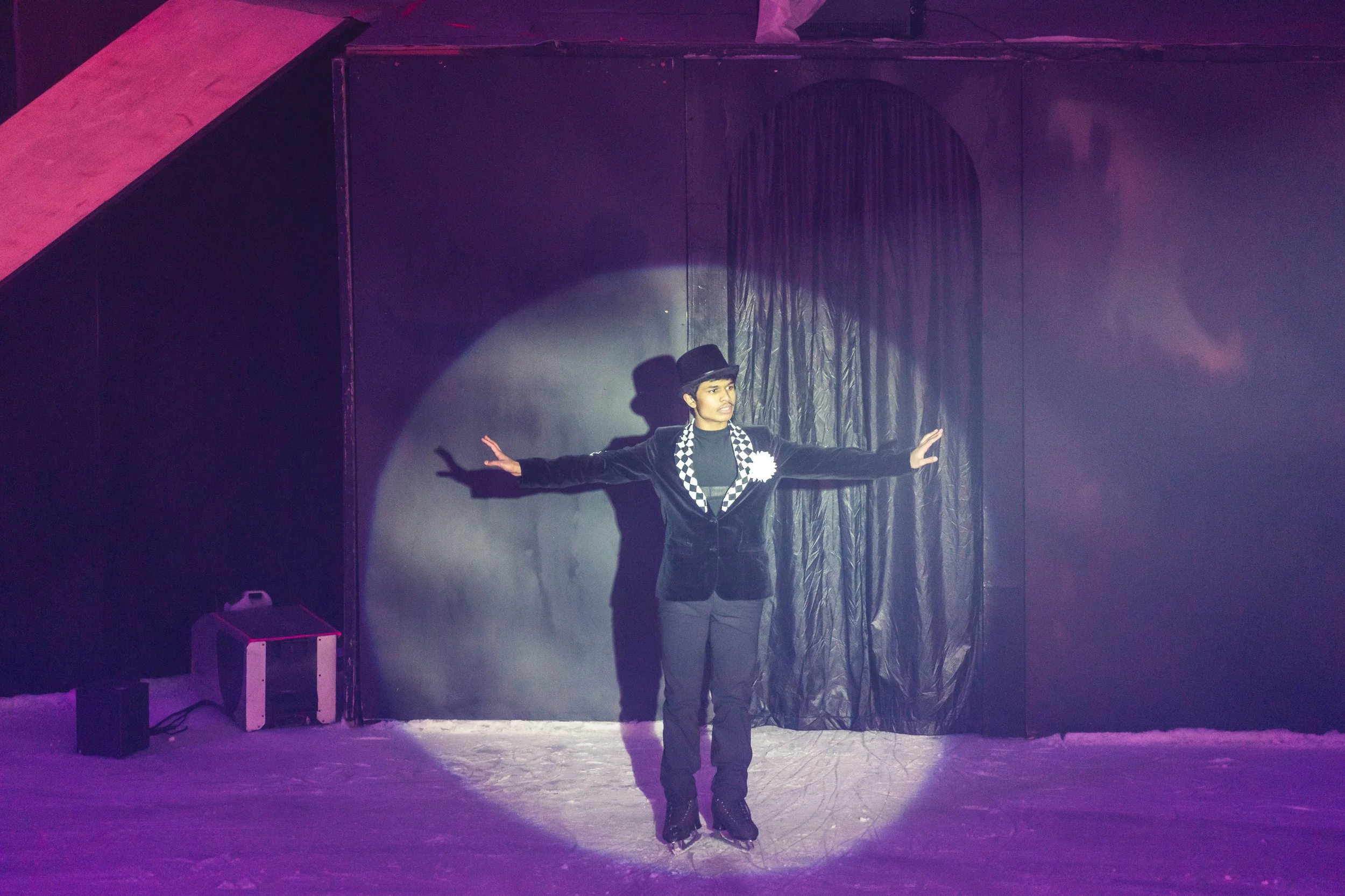 A young performer in black attire, top hat, and checkered scarf, standing on ice skating rink with arms outstretched, under stage lighting, in front of black curtains.