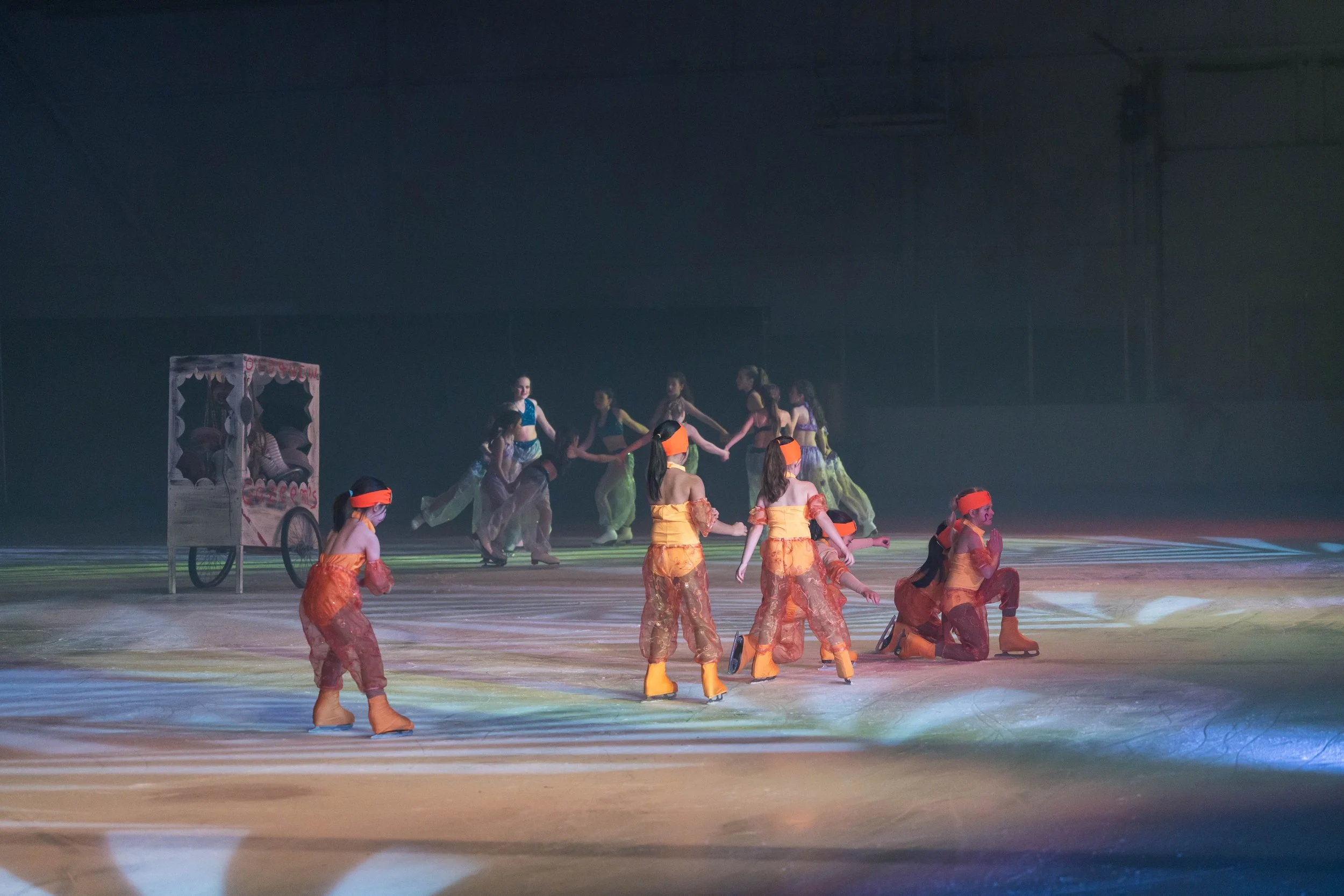 A group of young children in orange costumes and black masks with a dance troupe performing on stage, with some wearing colorful costumes, and a cart with a decorative top in the background.