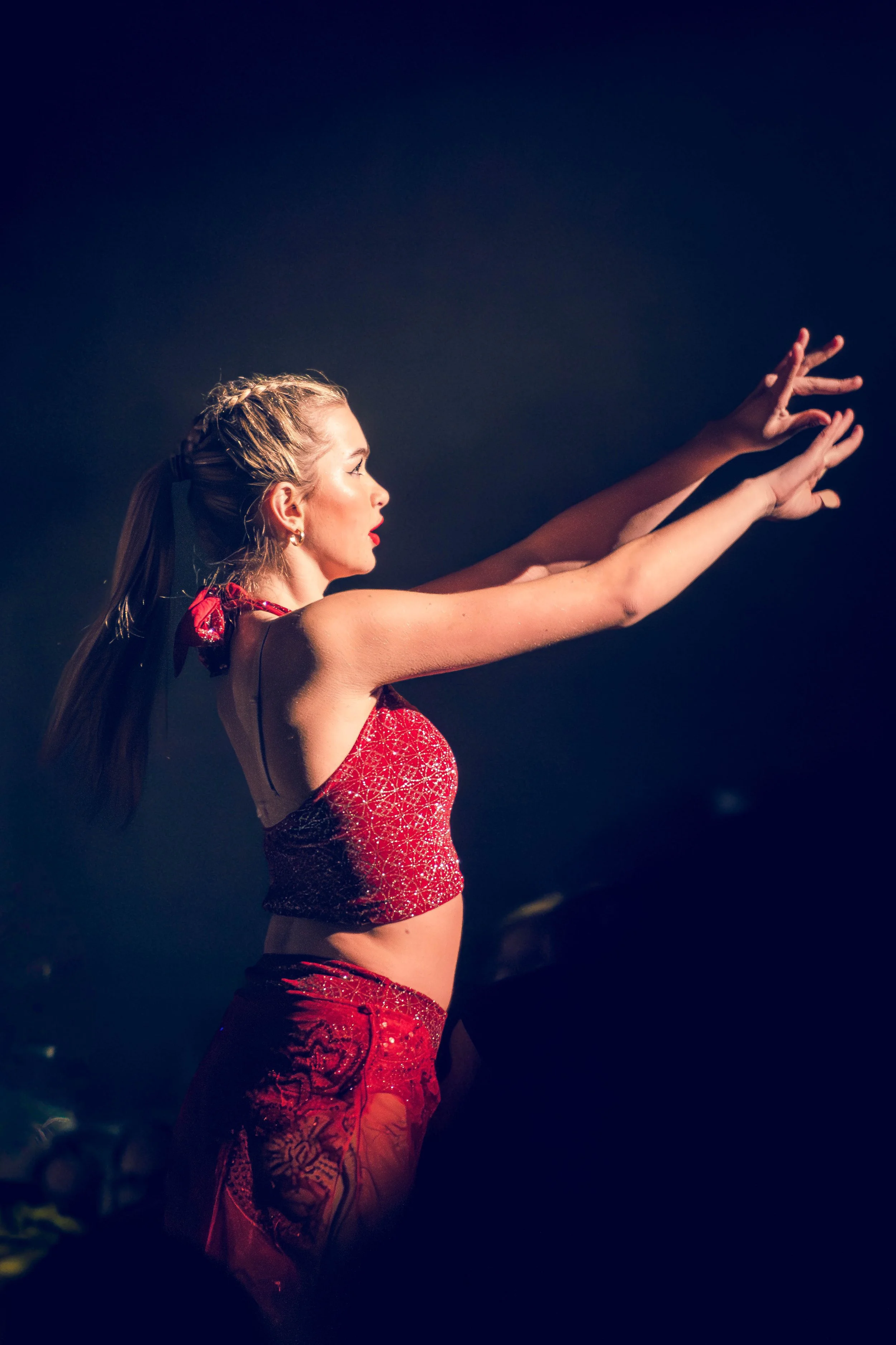 A woman with blonde hair performs a dance move on stage, wearing a red sequined top and matching pants. She has her arms extended forward and her hair styled in braids.