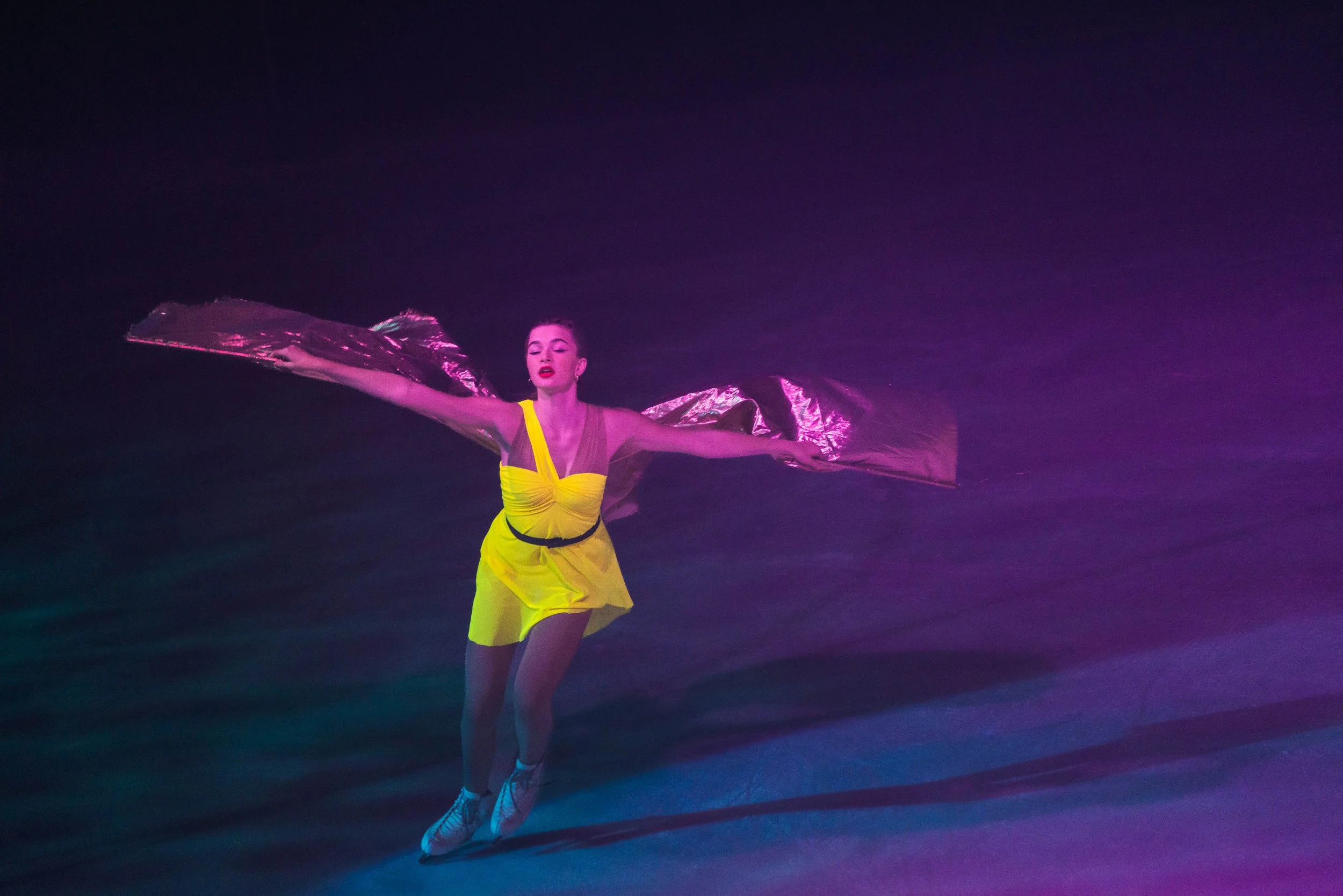 A woman in a yellow dress and sneakers dancing with metallic fabric flowing around her, under colorful stage lighting.