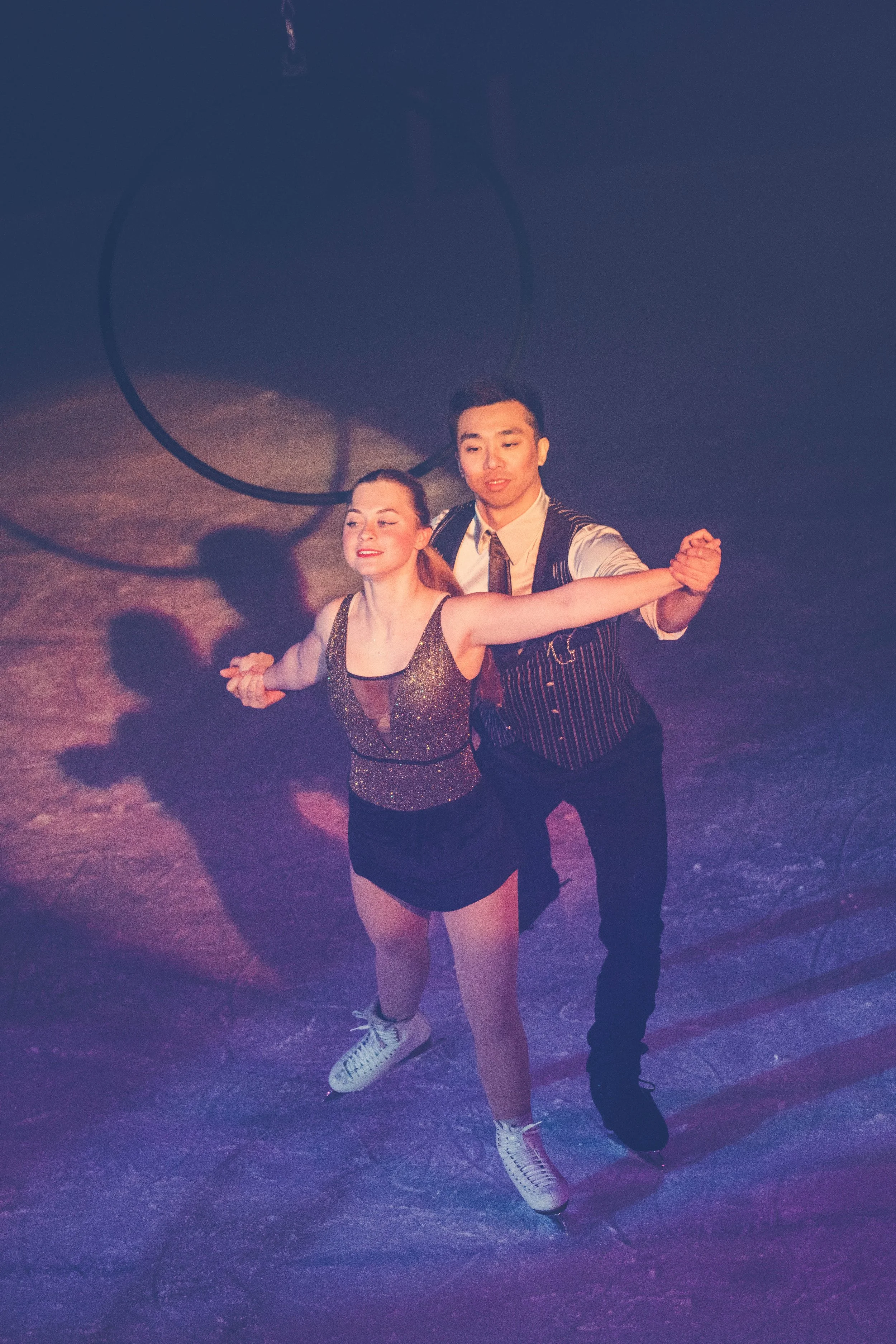 A man and woman ice skating on an ice rink, with the woman in front and the man holding her hand behind her.