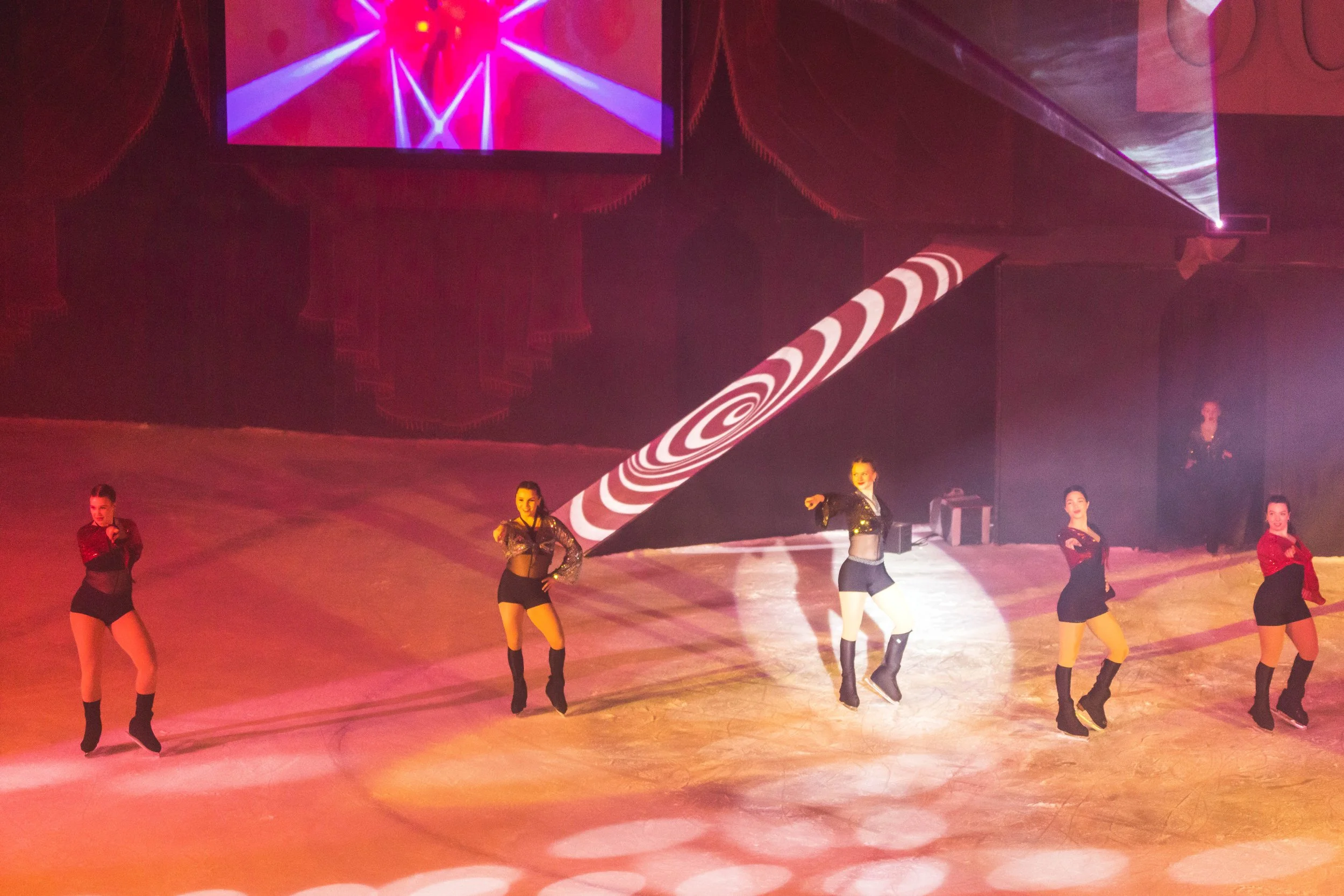 Stage performance with dancers in black and red costumes, colorful lighting, and a large screen displaying bright lights and patterns.