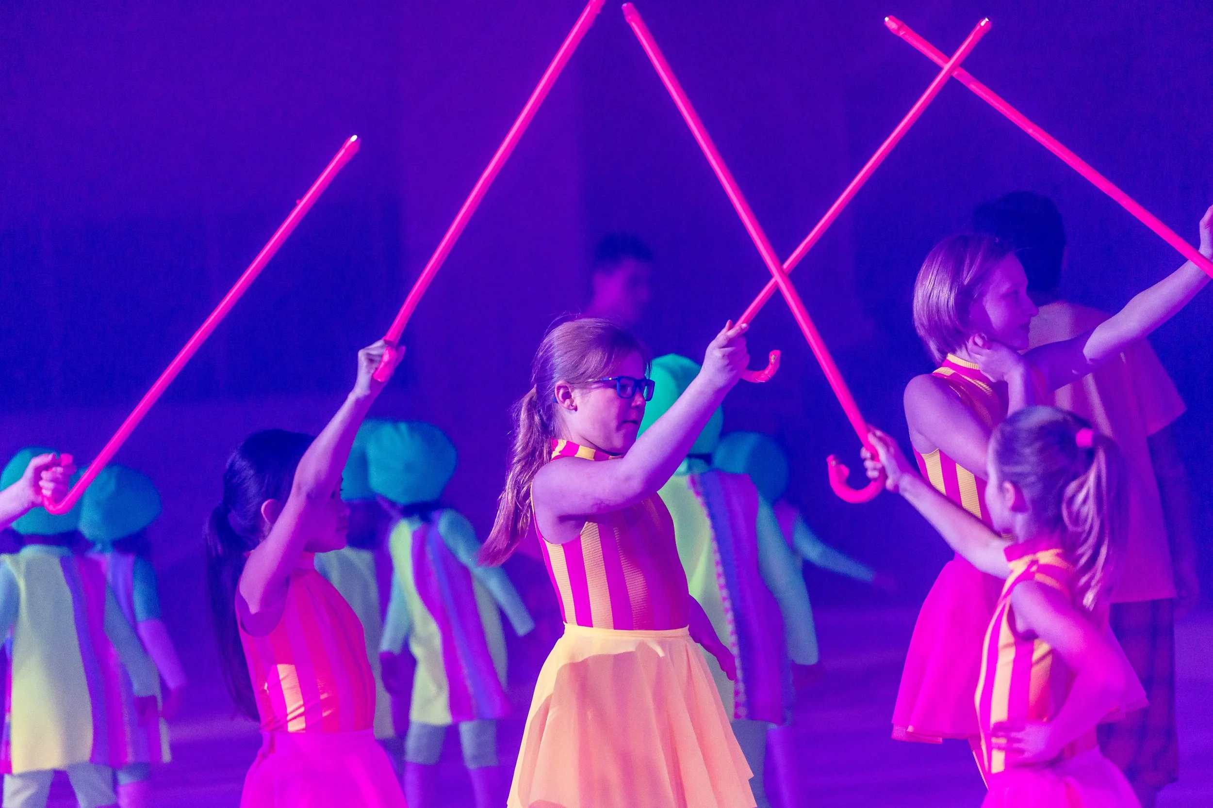 A group of children dressed in colorful costumes performing a dance with pink glowing sticks in a dark, neon-lit setting.