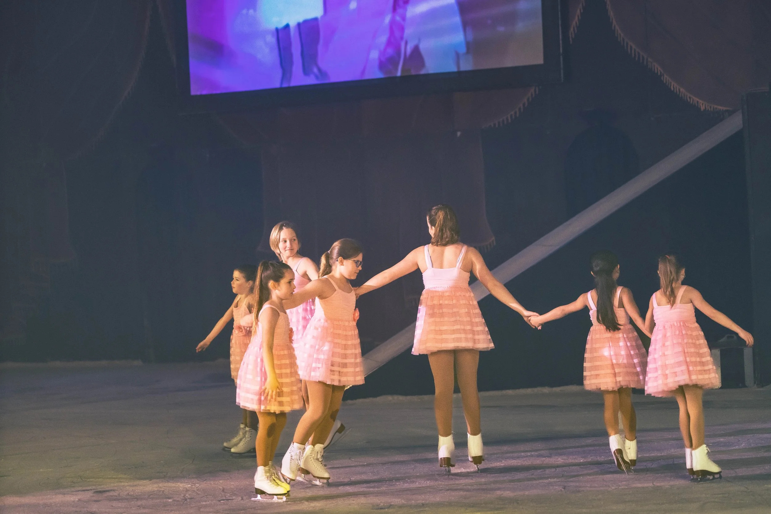A group of girls dressed in pink costumes with plaid skirts and skating on an indoor ice rink, holding hands and performing a synchronized dance.