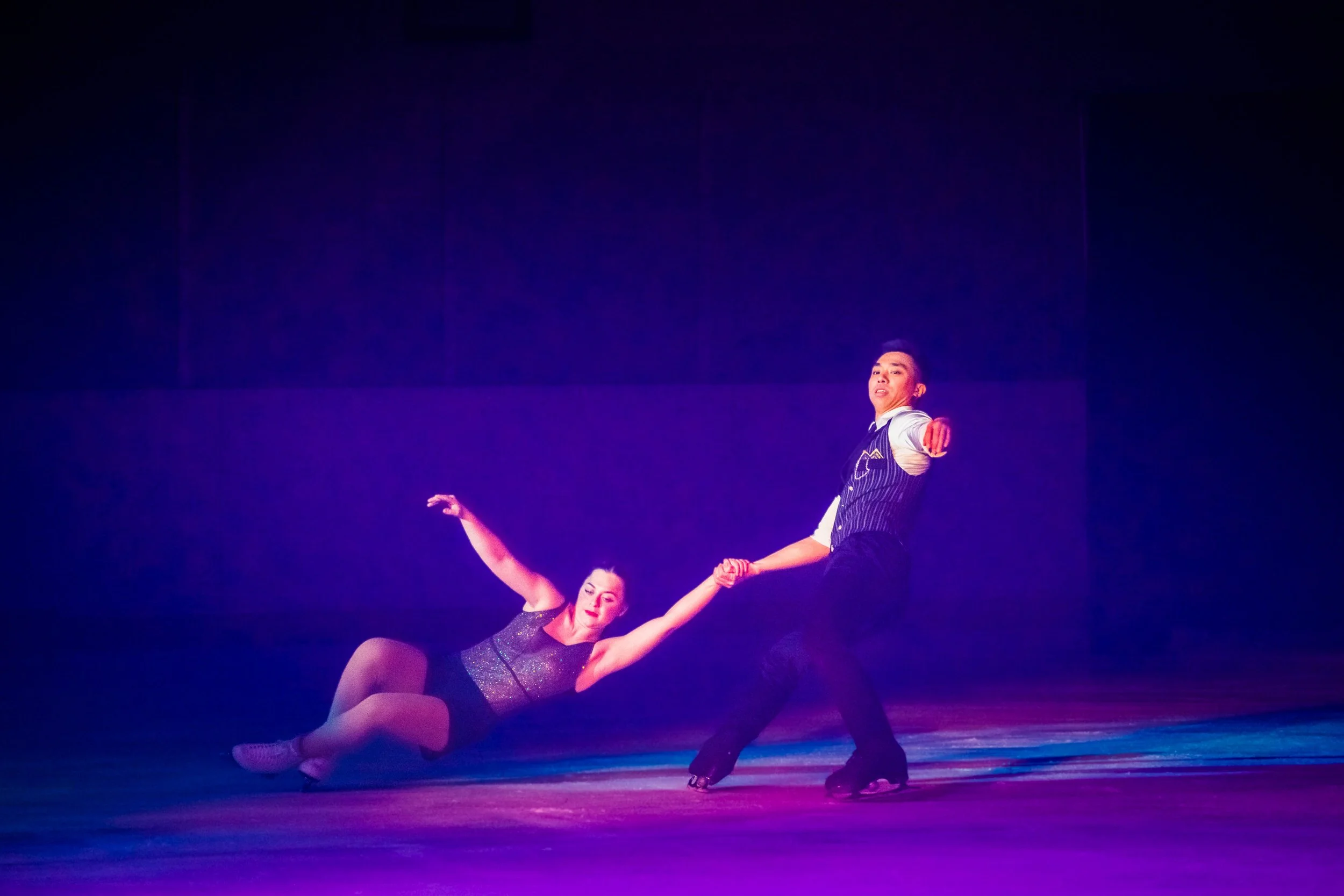 Two dancers performing on stage under purple lighting, with one dancer lying on the floor extending her leg upward and the other dancer standing beside her holding her hand.