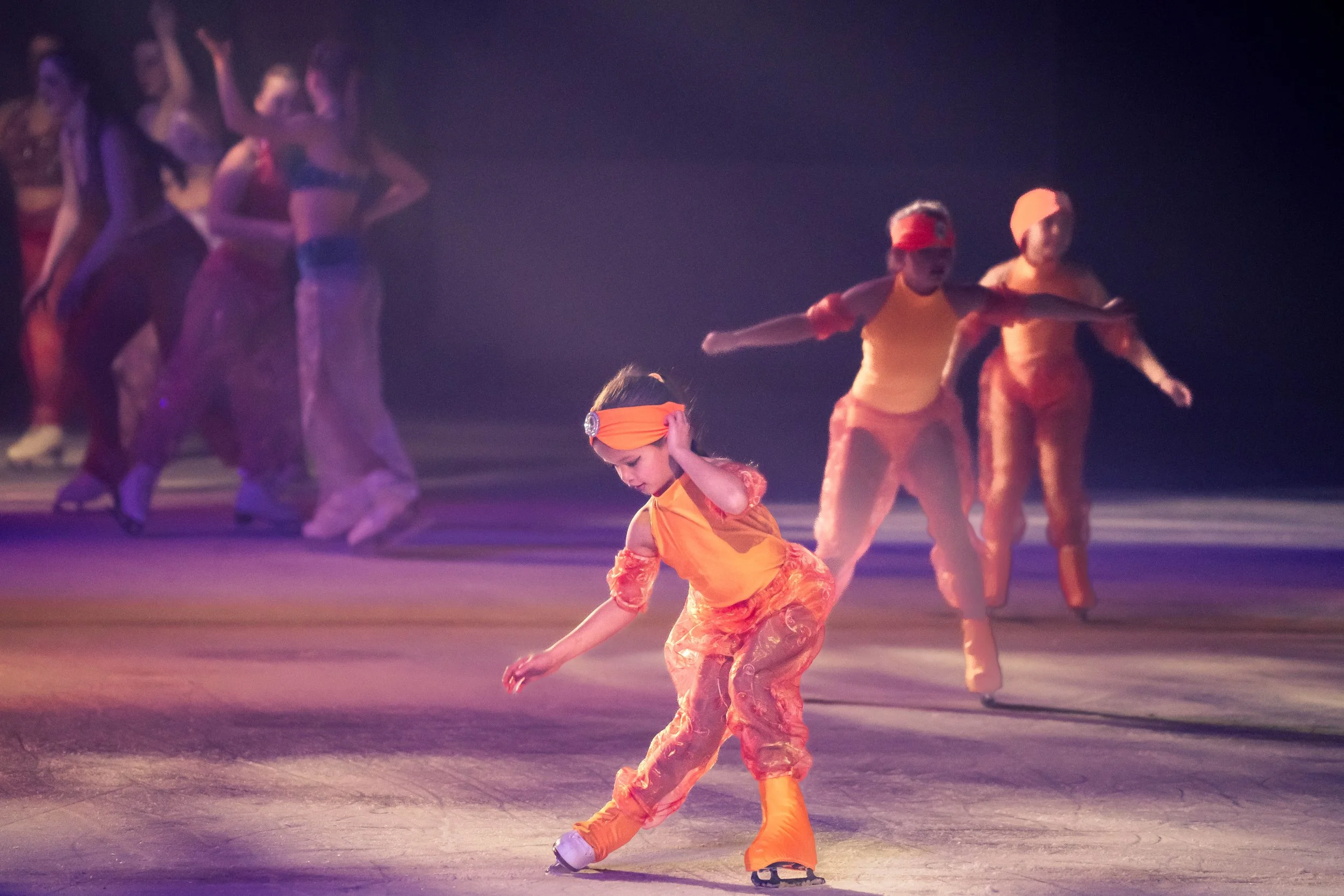 Children in colorful costumes performing a dance routine on stage with purple and pink lighting.