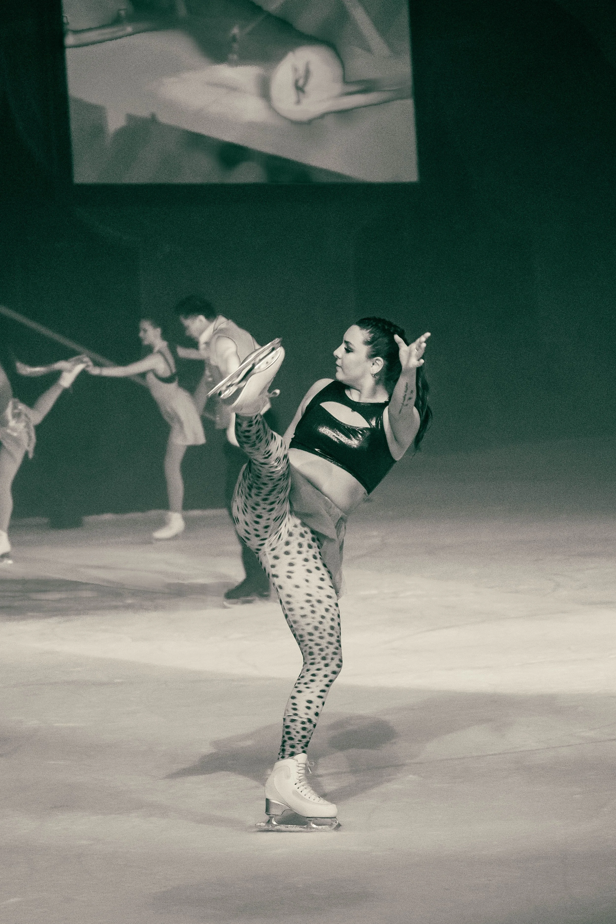 A female figure skater in a black and white outfit performing a move on ice skates during a dance routine, with other skaters in the background.