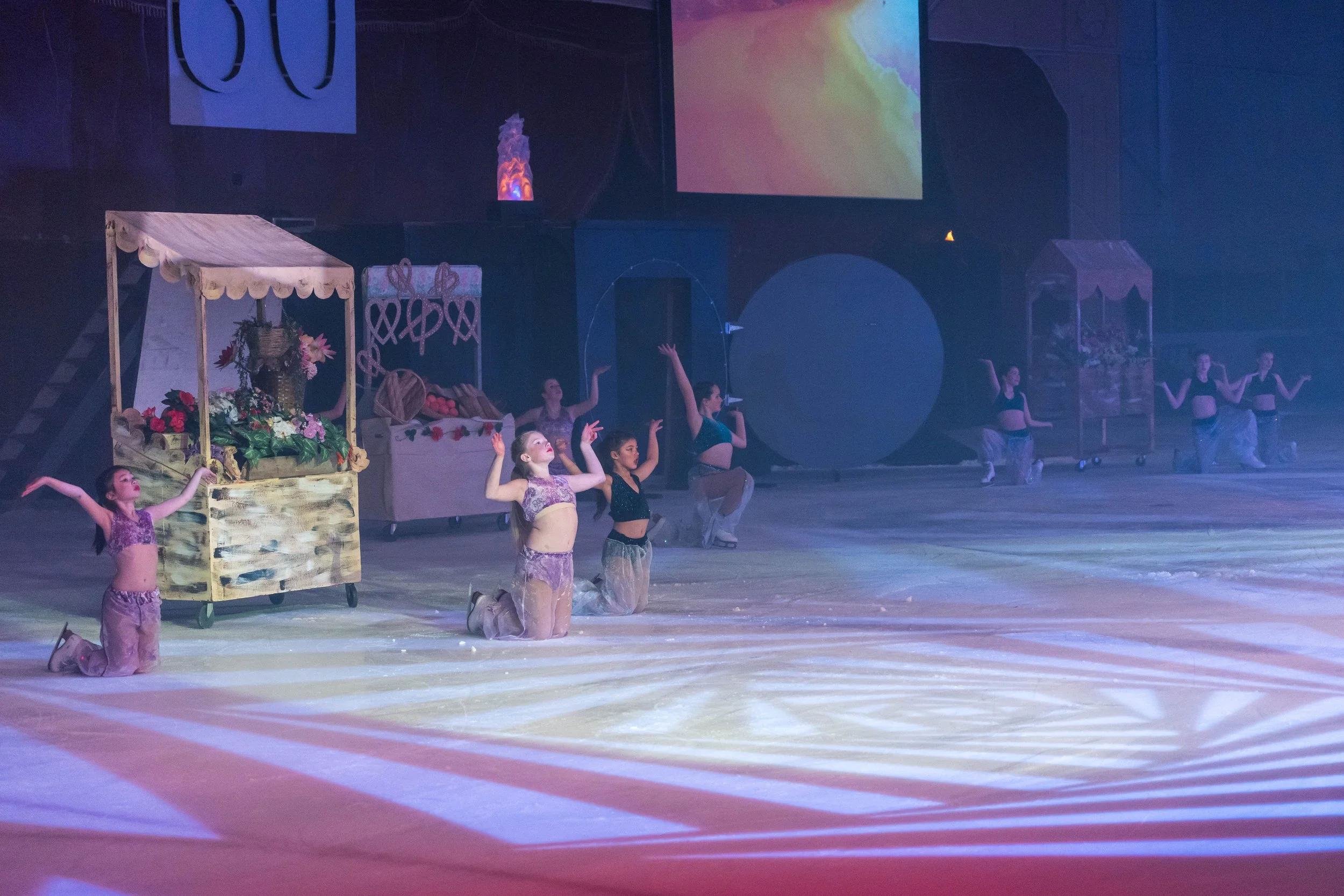 Young female figure skaters wearing costumes rehearsing or performing on ice in a theater with stage set pieces and large screens.